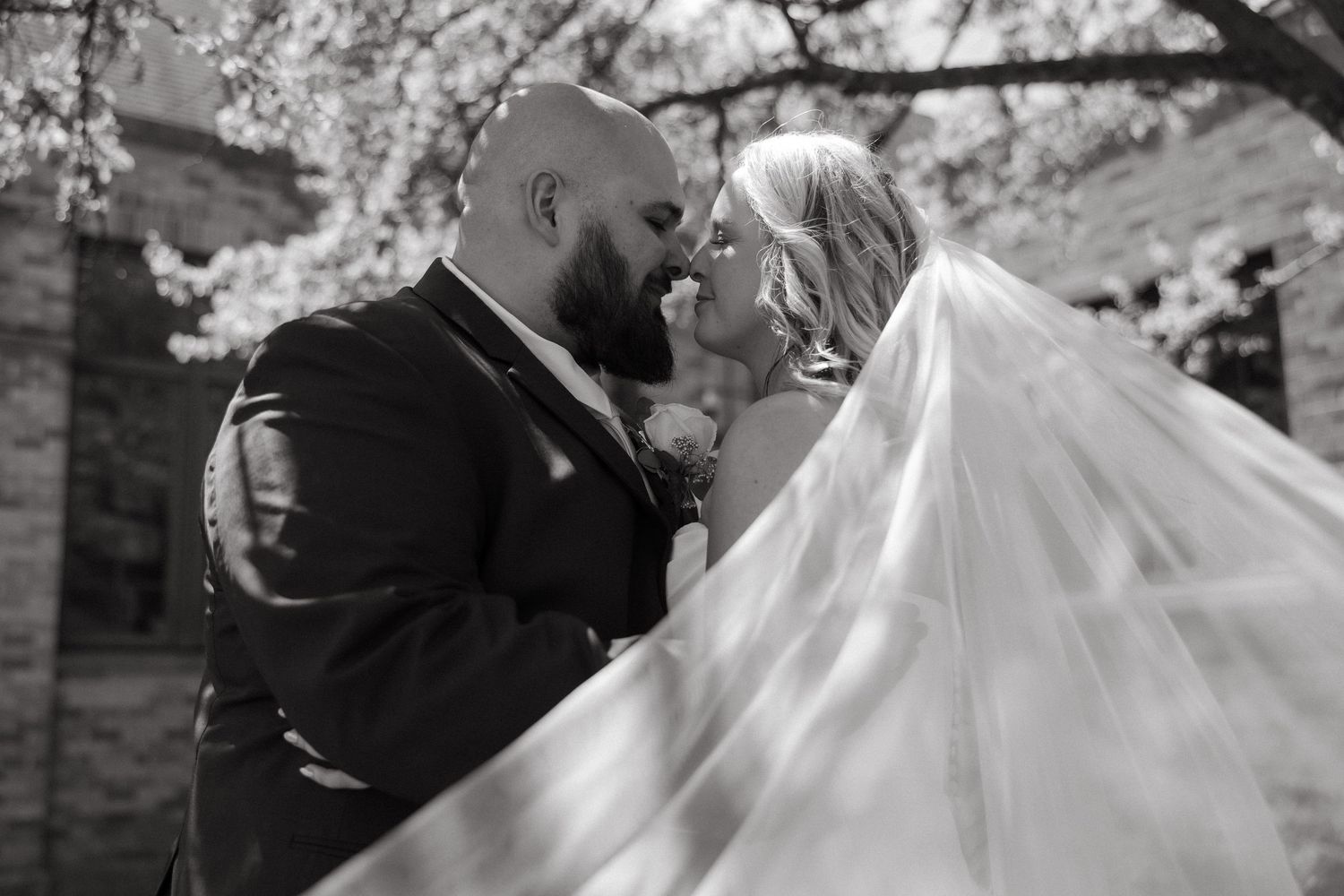 Black and white wedding photo of bride and groom nearly kissing under blooming trees, veil flowing dramatically.