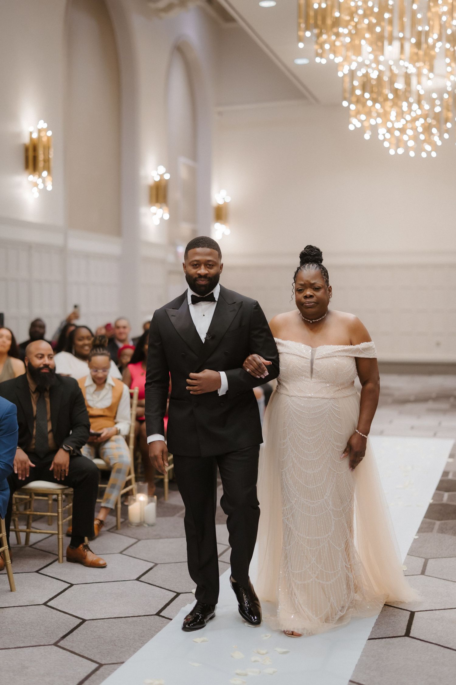 Groom in black tuxedo escorts bride in off-shoulder lace gown down white aisle at elegant wedding.