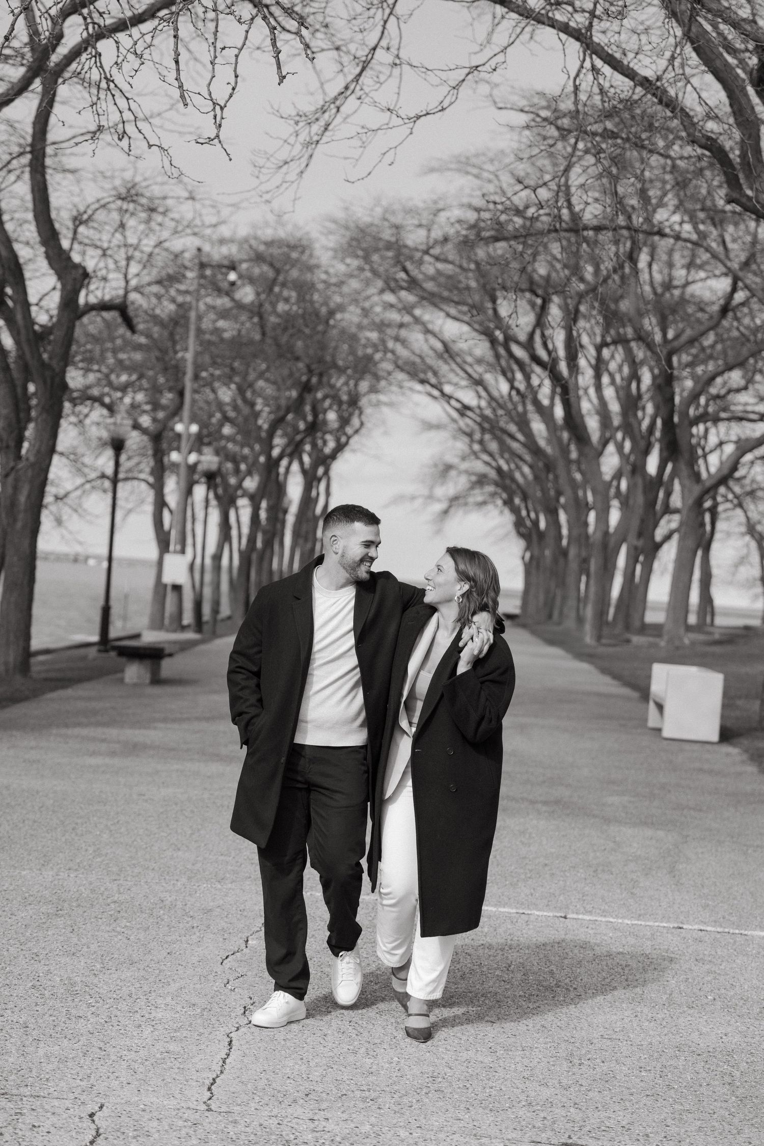 Black and white photo of a couple walking and smiling at each other on a tree-lined path.