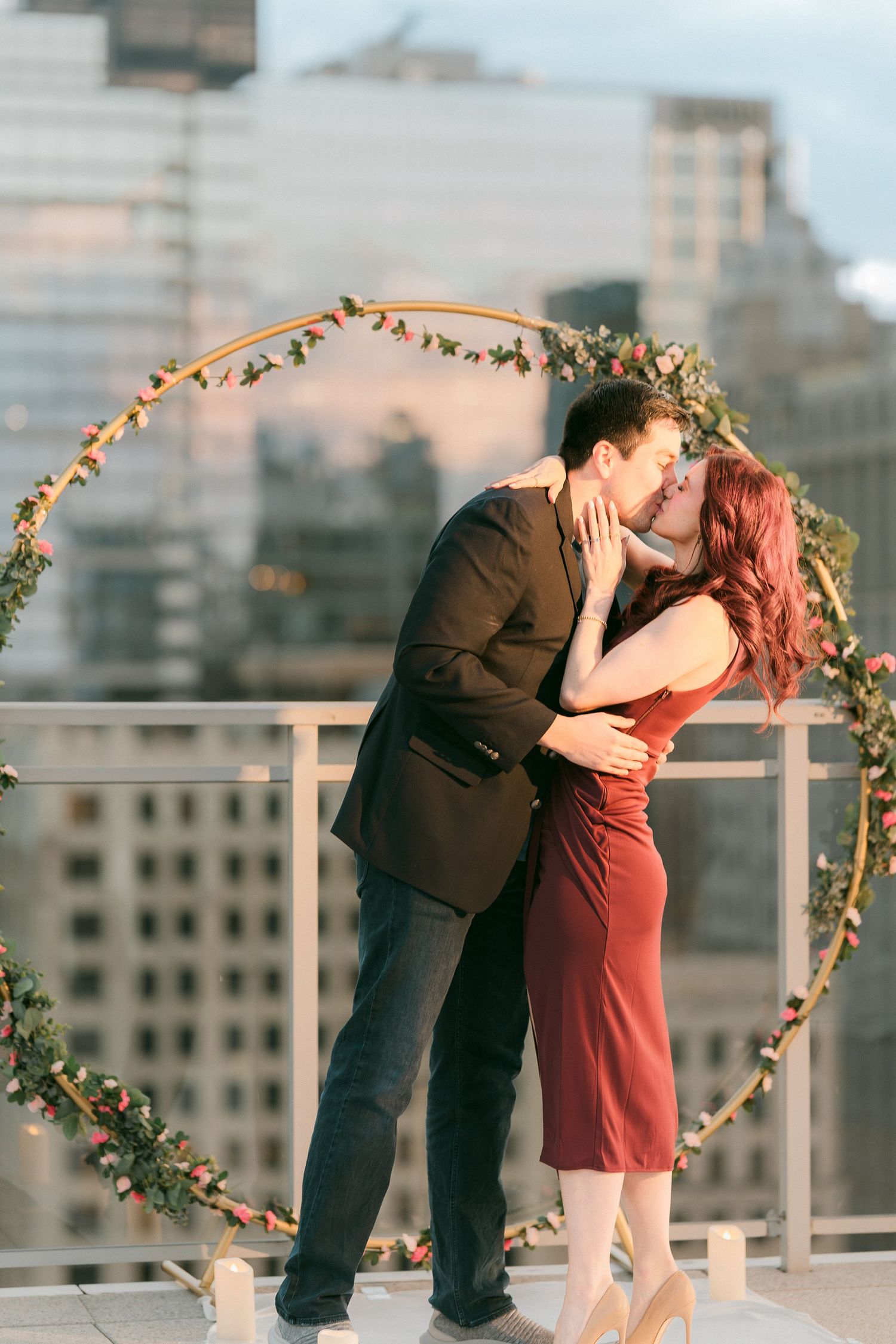 Couple kisses romantically in front of circular floral arch on rooftop with Chicago city skyline at dusk.