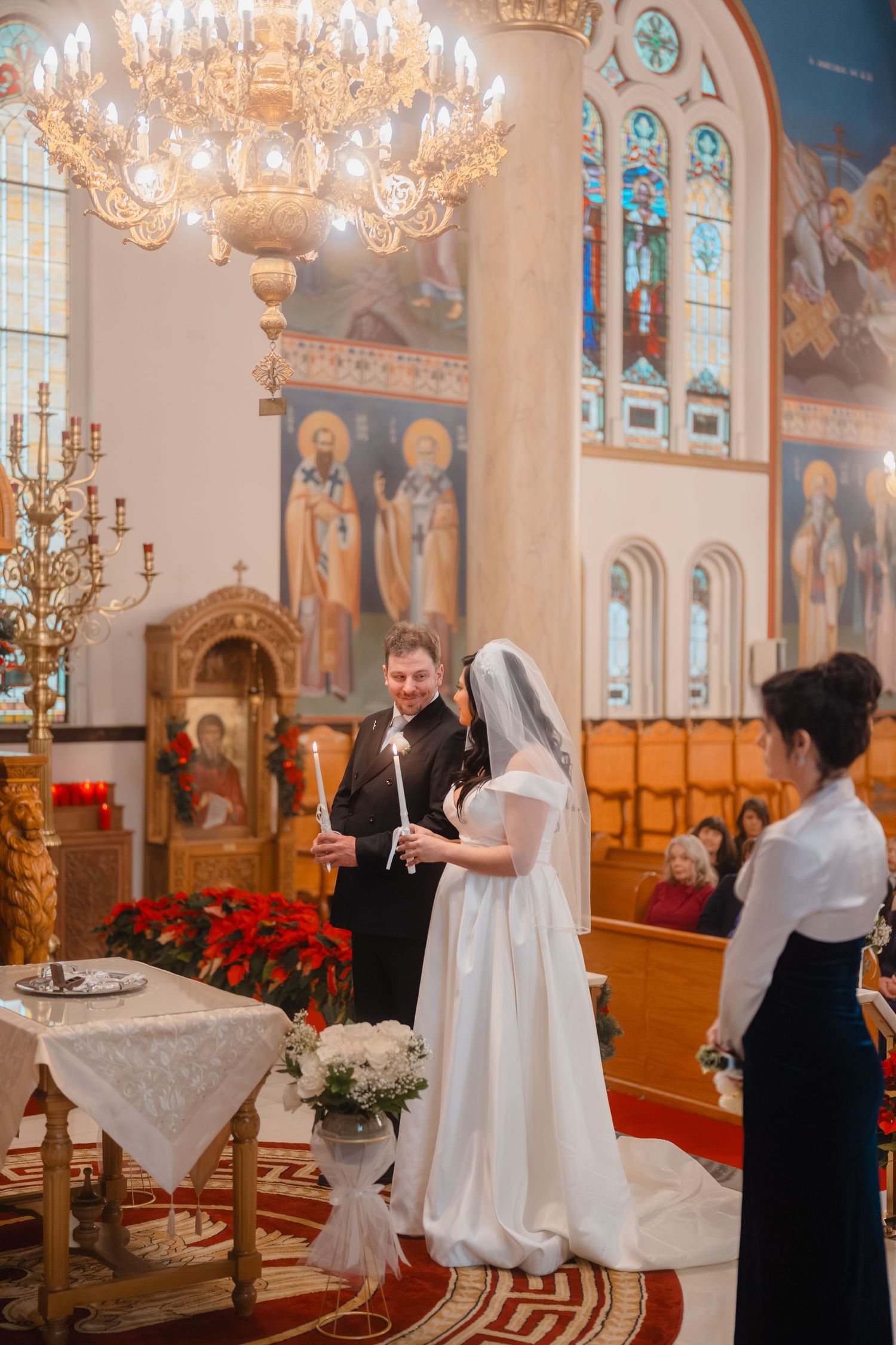 Bride and groom hold candles during Orthodox wedding ceremony beneath golden chandelier with colorful religious murals.