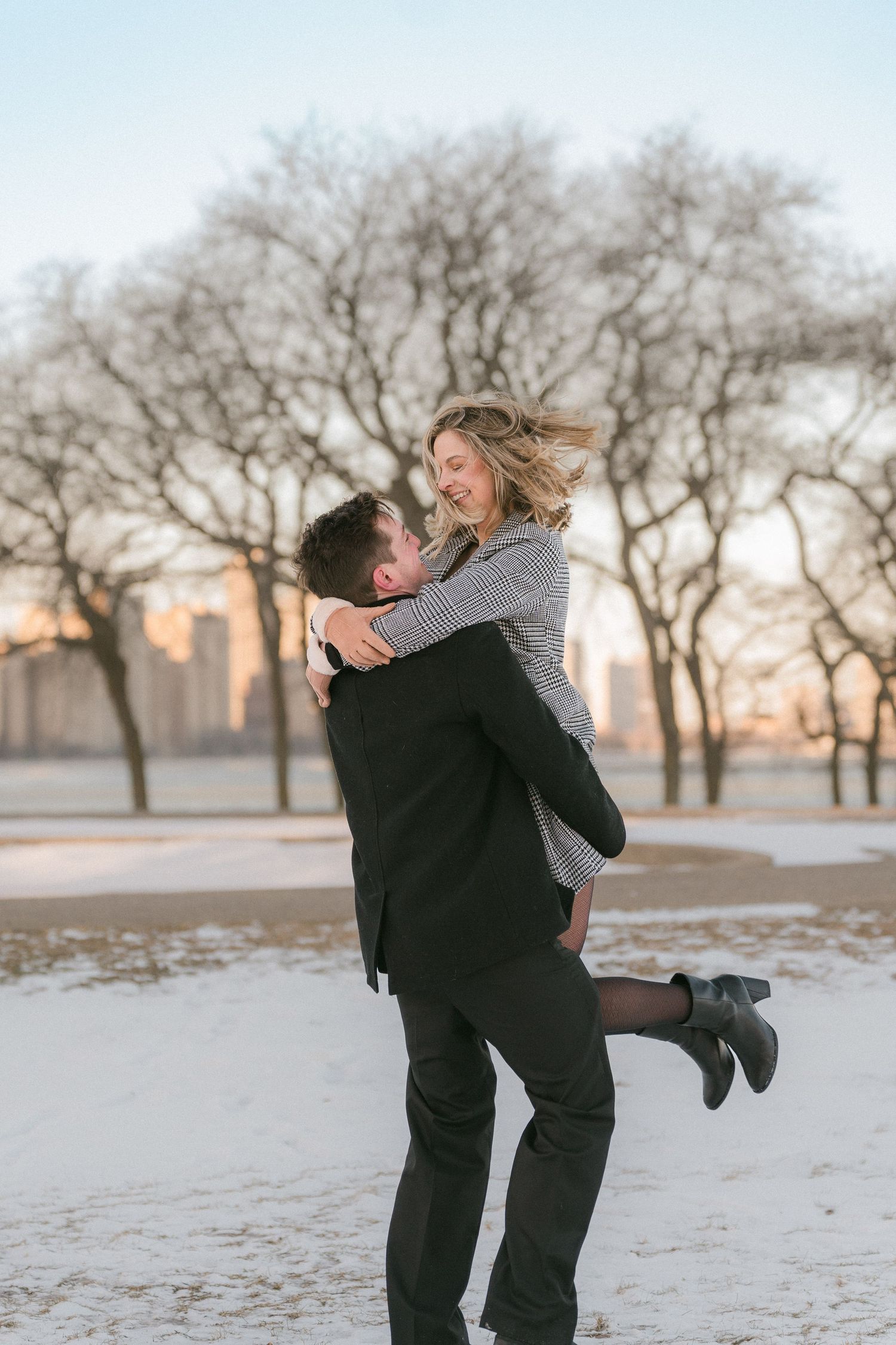 A man lifts and spins a smiling woman in a snowy park with bare trees at golden hour.