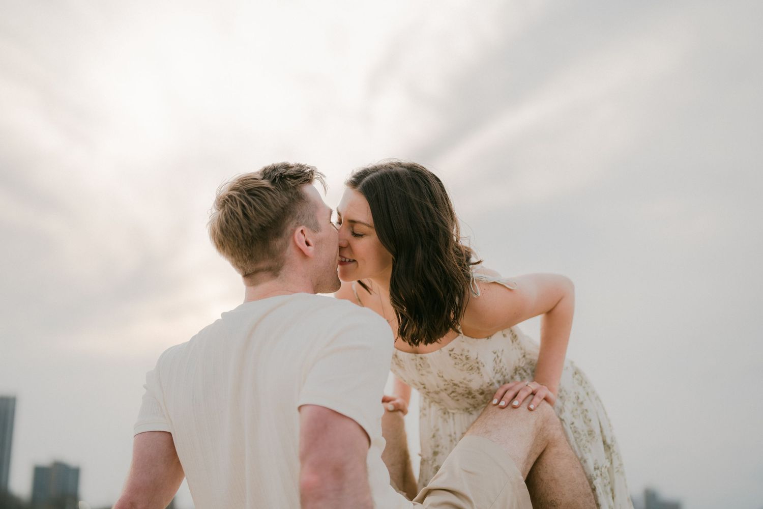 Couple sharing a romantic kiss against a dramatic cloudy sky with a city skyline visible in the background.