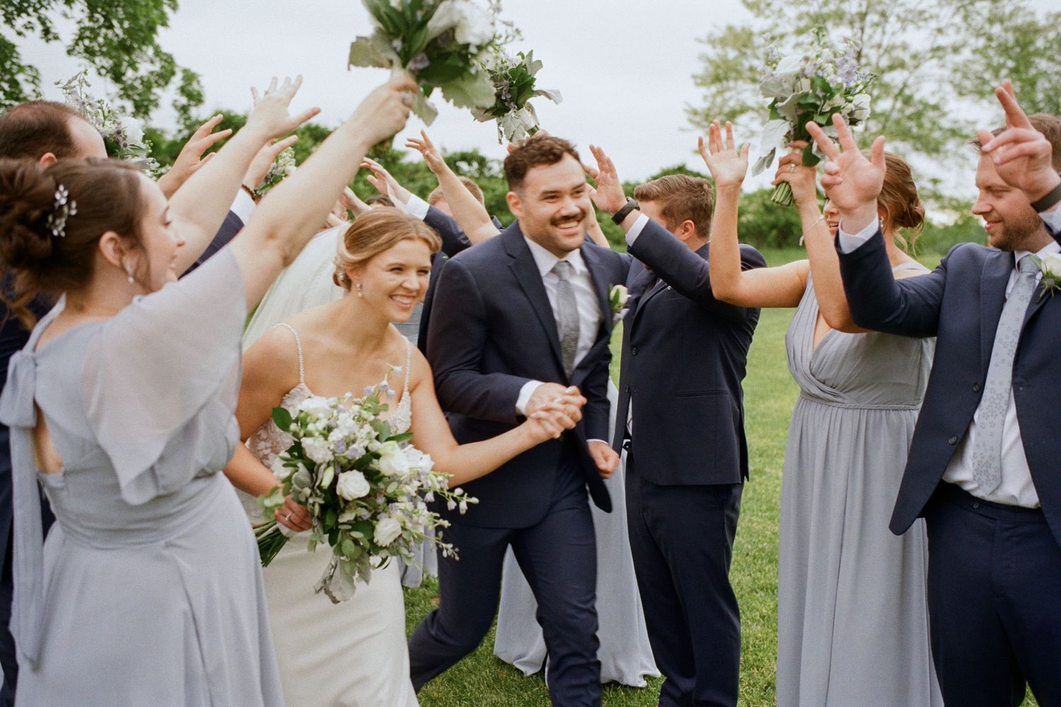 Joyful bride and groom celebrate with wedding party in blue-gray attire outdoors, raising hands in celebration.