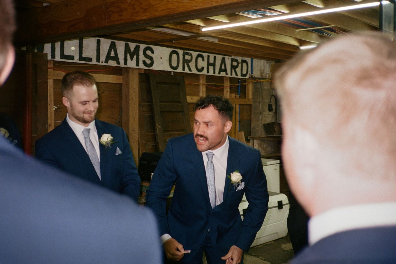 Groomsmen in navy suits with boutonnieres gather inside Williams Orchard barn venue before a wedding ceremony.