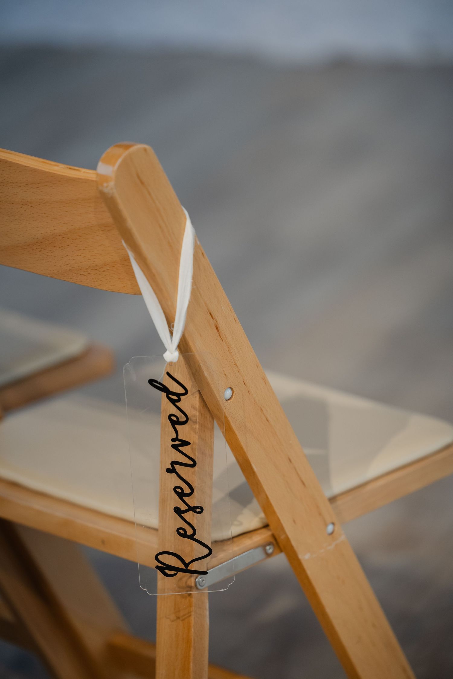 A wooden folding chair with a clear acrylic reserved sign hanging from white ribbon at an outdoor wedding ceremony.