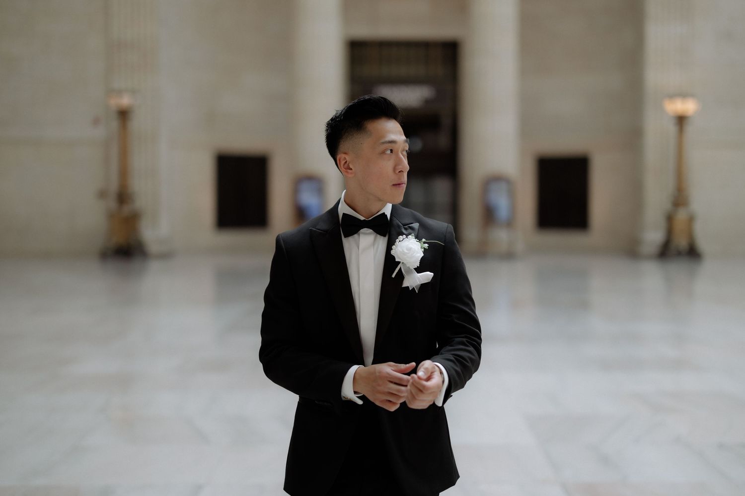 Groom in black tuxedo with white boutonniere stands in grand marble wedding venue hall.