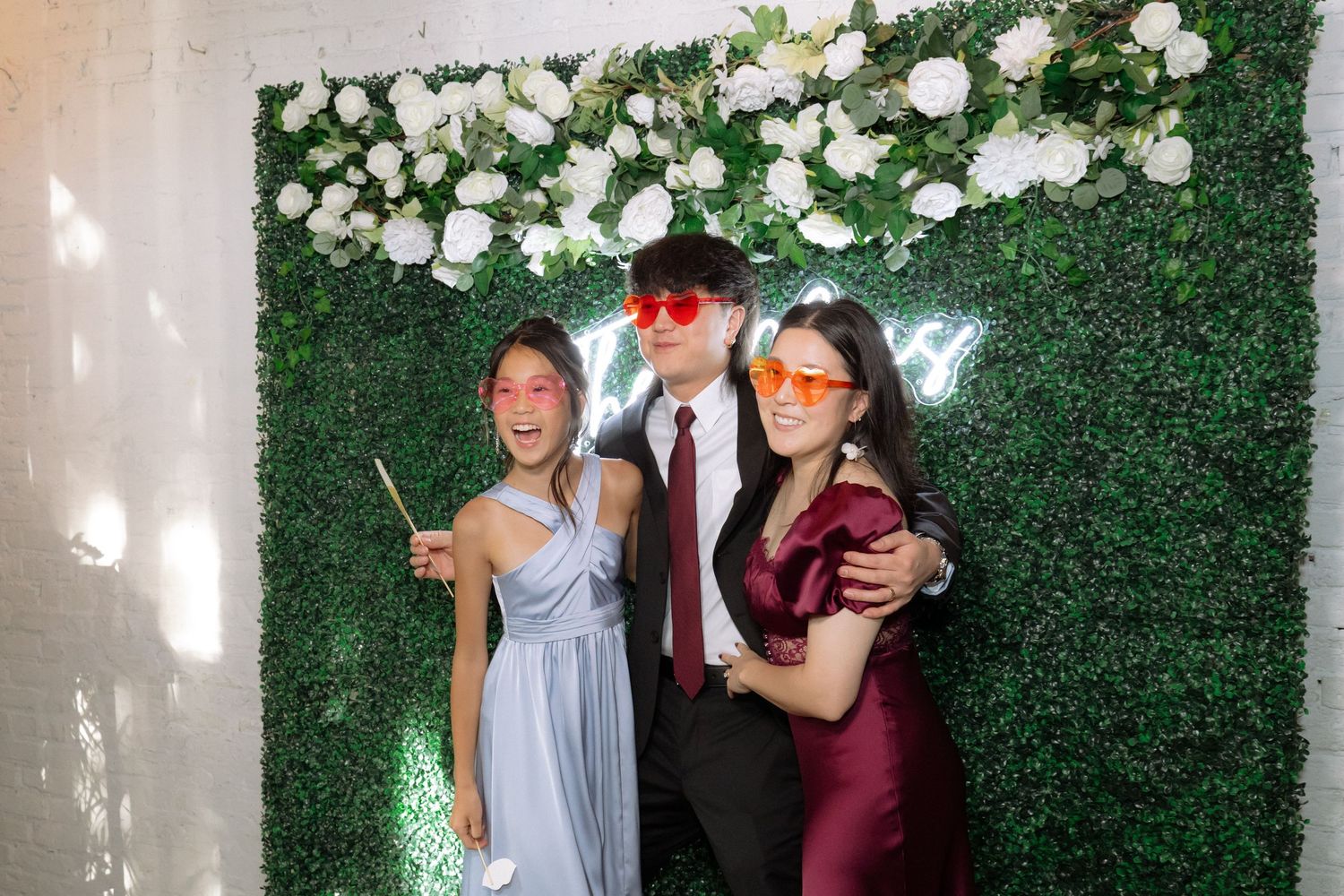 Three young people posing at a wedding photo booth with green hedge wall and white floral backdrop.