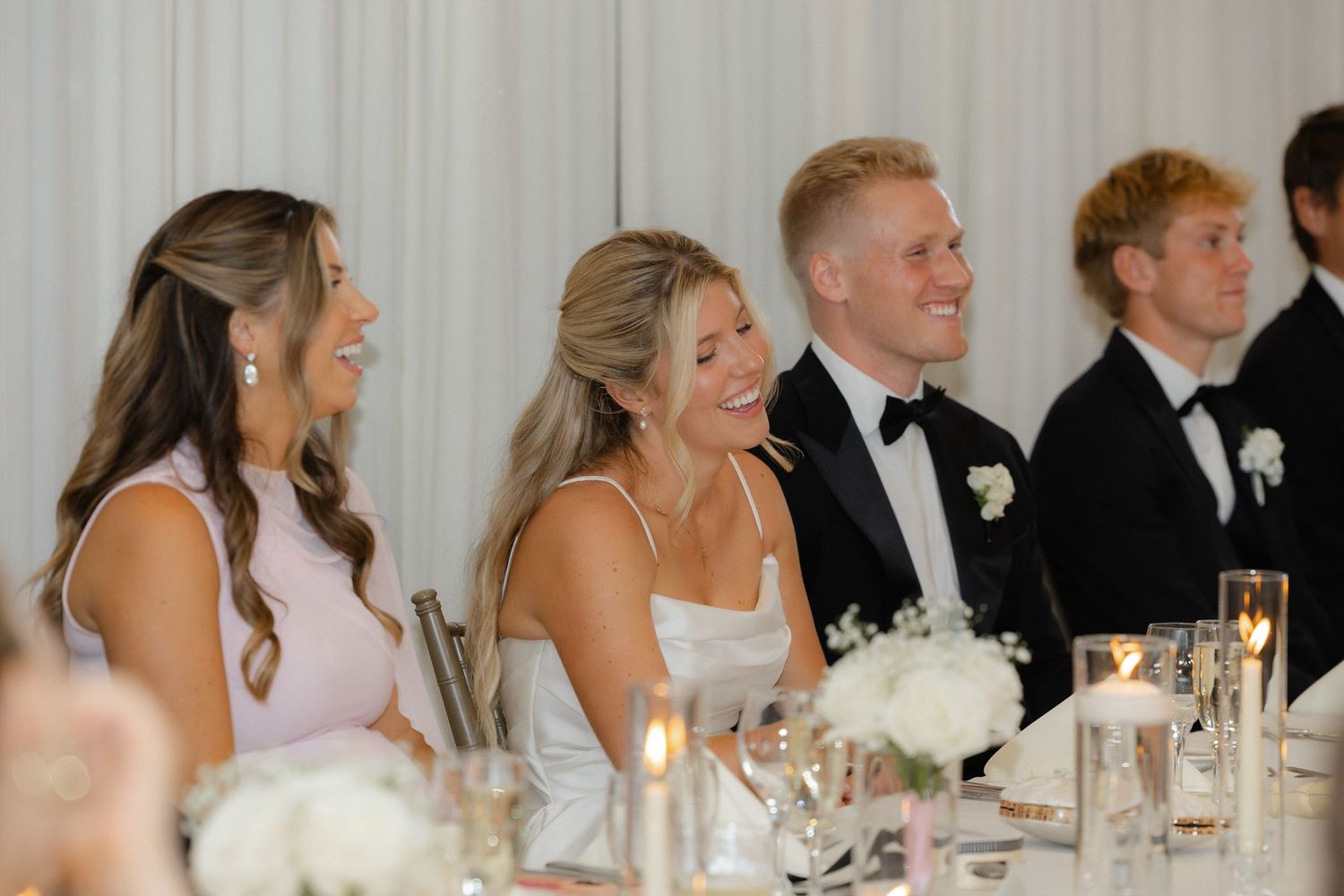 Bride and wedding party laugh joyfully at the head table during elegant white wedding reception.