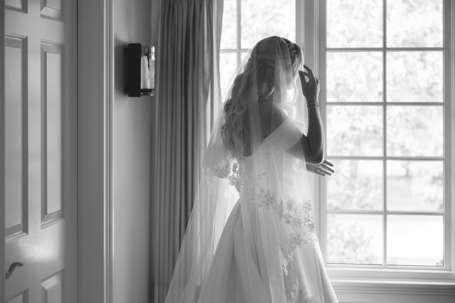 Black and white silhouette of a bride in a full wedding gown and veil standing by a window.