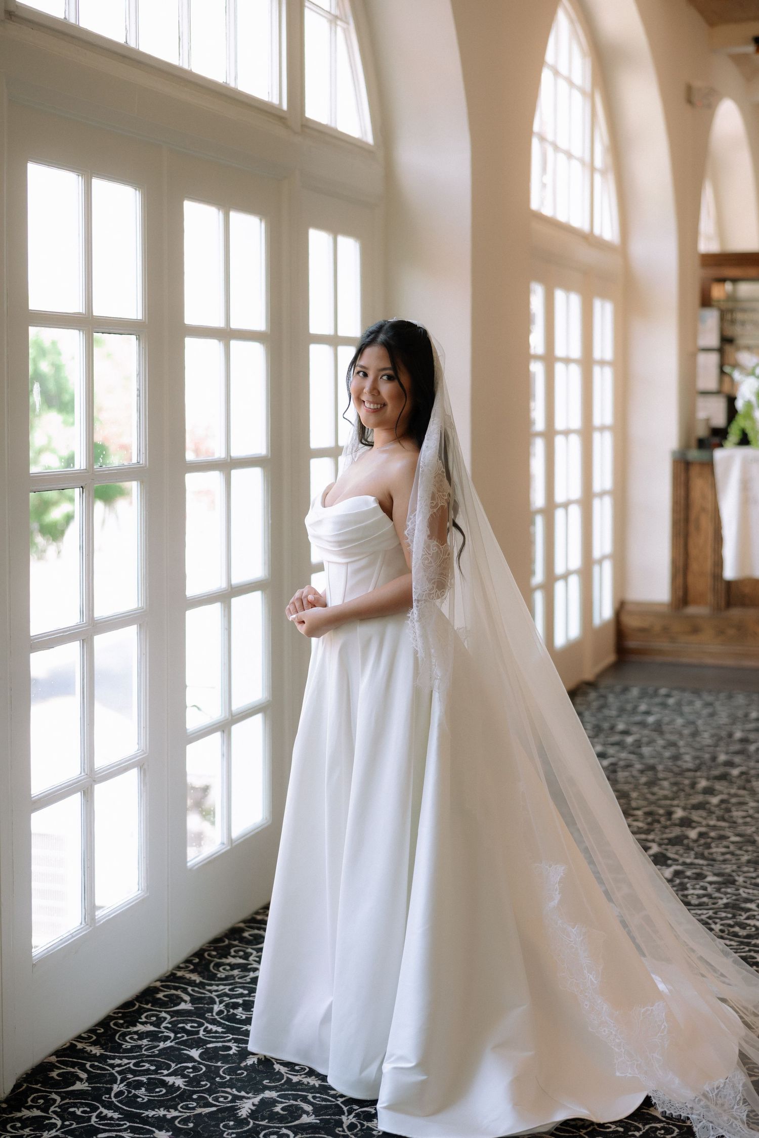 Elegant bride in strapless white gown with cathedral veil poses by tall windows in bright hallway.