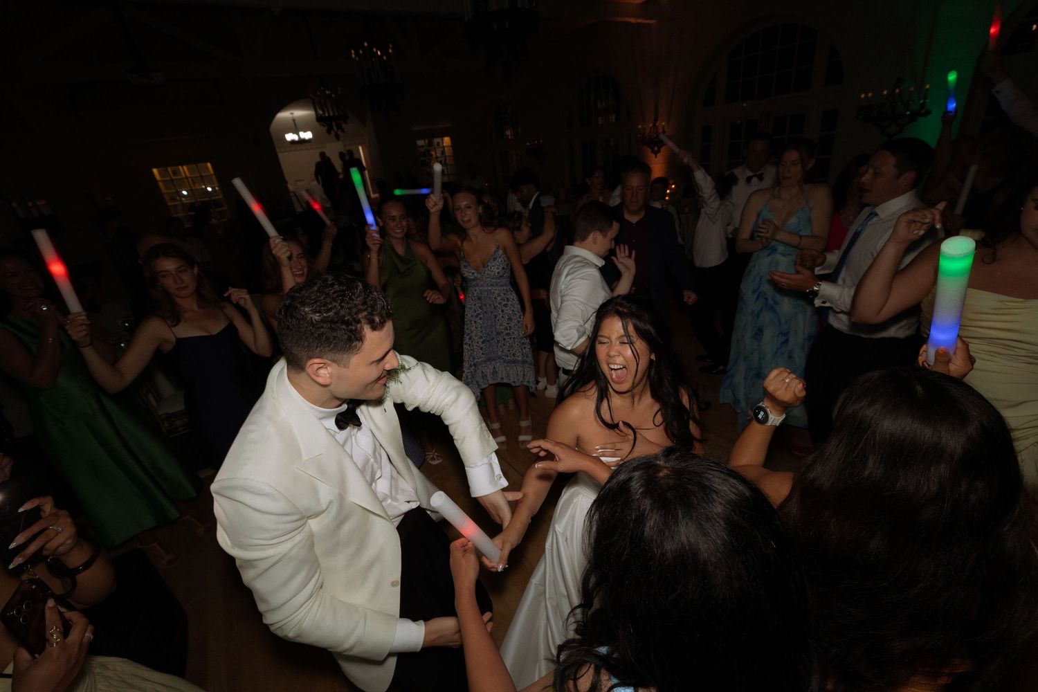 Bride and groom dance joyfully with guests holding glow sticks at a lively wedding reception.