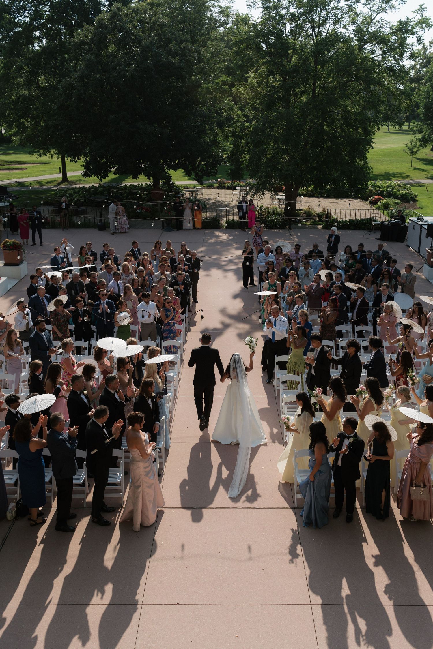 Aerial view of bride and groom walking back down the aisle after wedding ceremony with cheering guests holding parasols.
