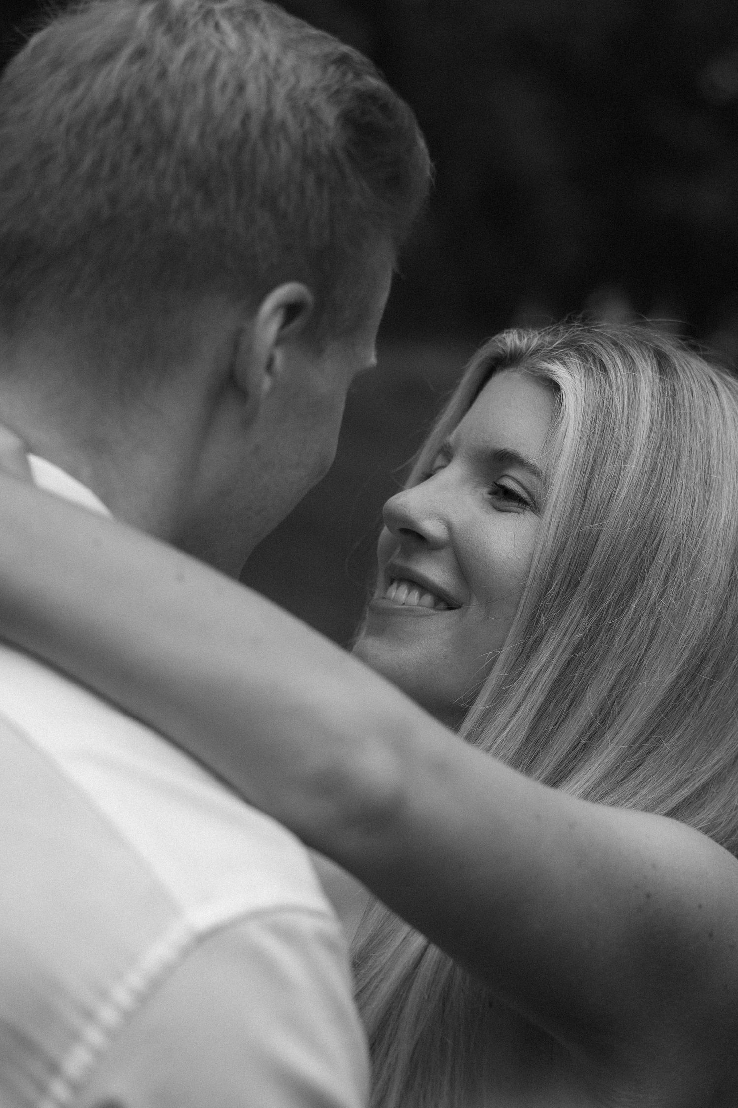 Black and white photo of a smiling blonde woman embracing a man outdoors, gazing lovingly at each other.