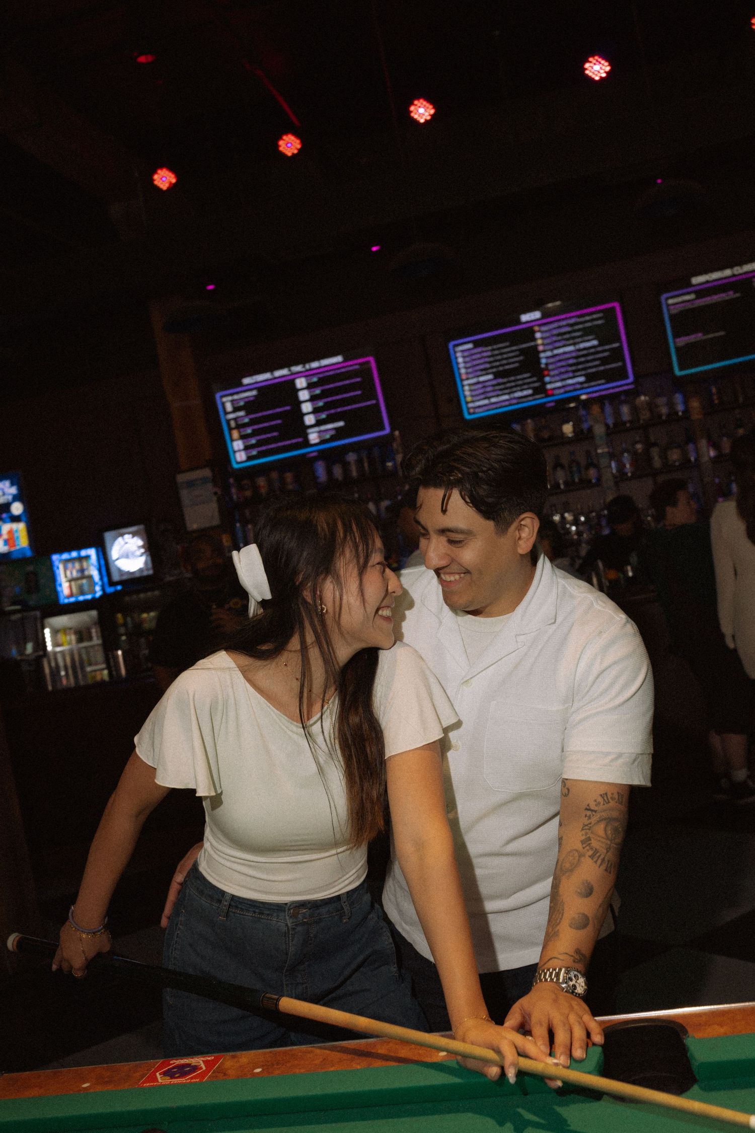 Smiling couple leaning close together over a pool table in a dimly lit bar.