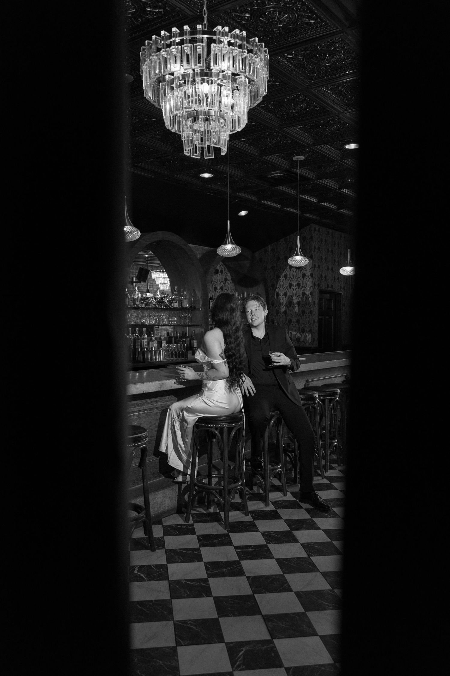 Black and white photo of a couple at an elegant bar with chandelier and checkered floor.