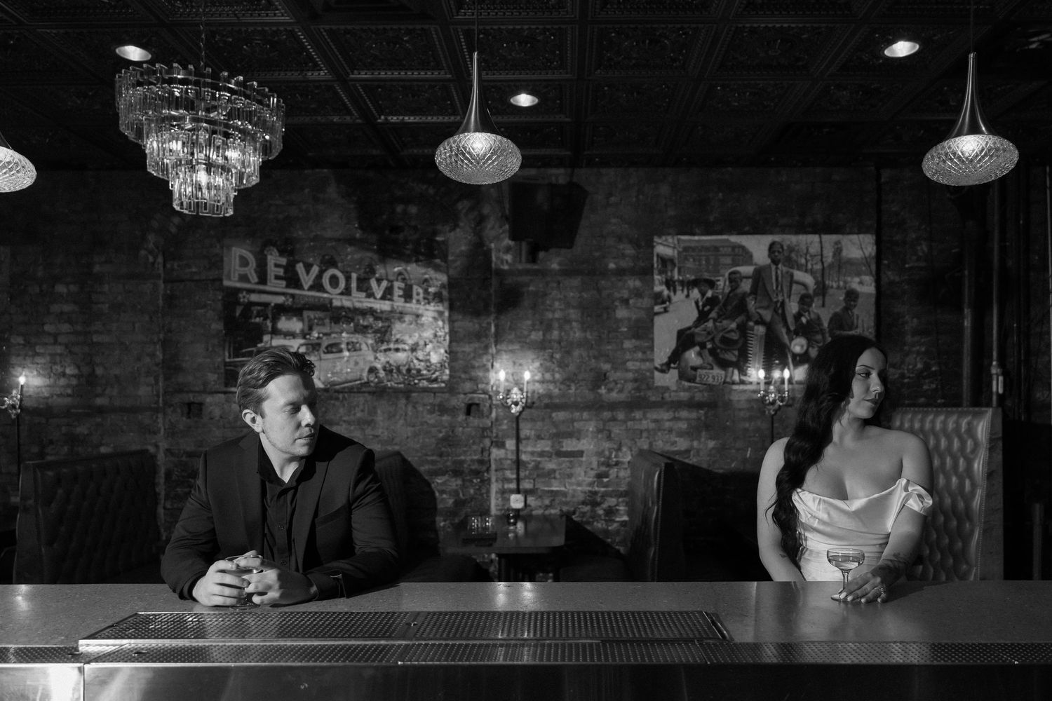 Black and white photo of bride and groom sitting apart at a bar in a rustic venue with chandelier.