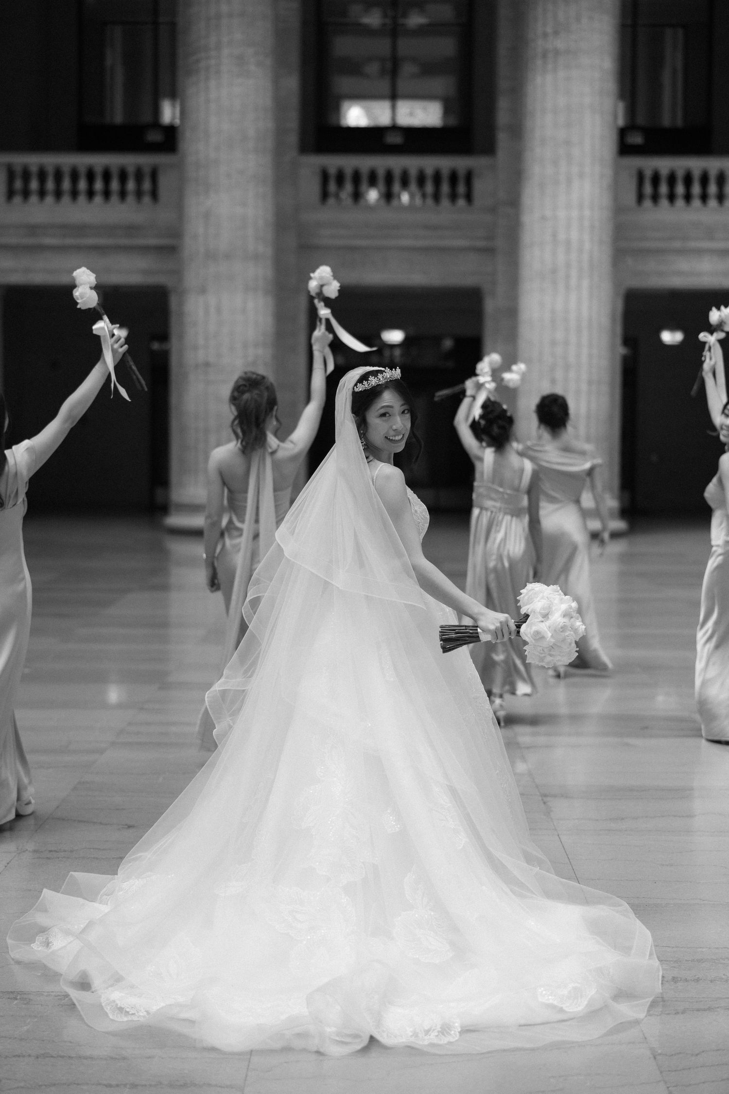Black and white photo of smiling bride in tiara and ballgown with bridesmaids raising bouquets in hall.
