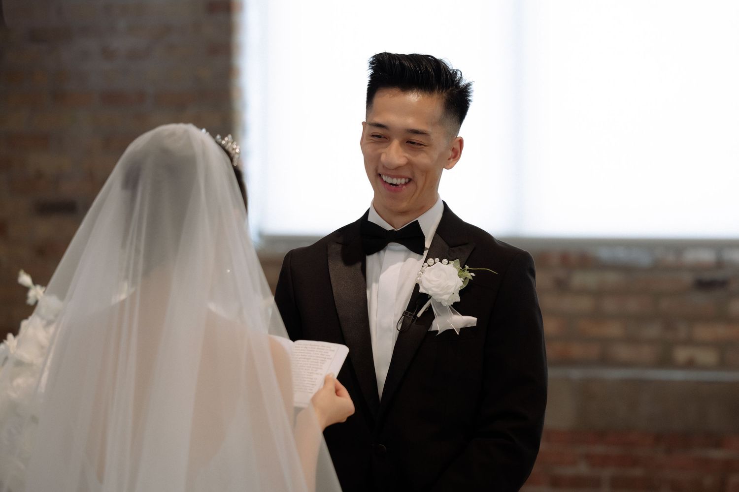 Groom in black tuxedo smiling at bride in white veil during wedding ceremony in brick venue.