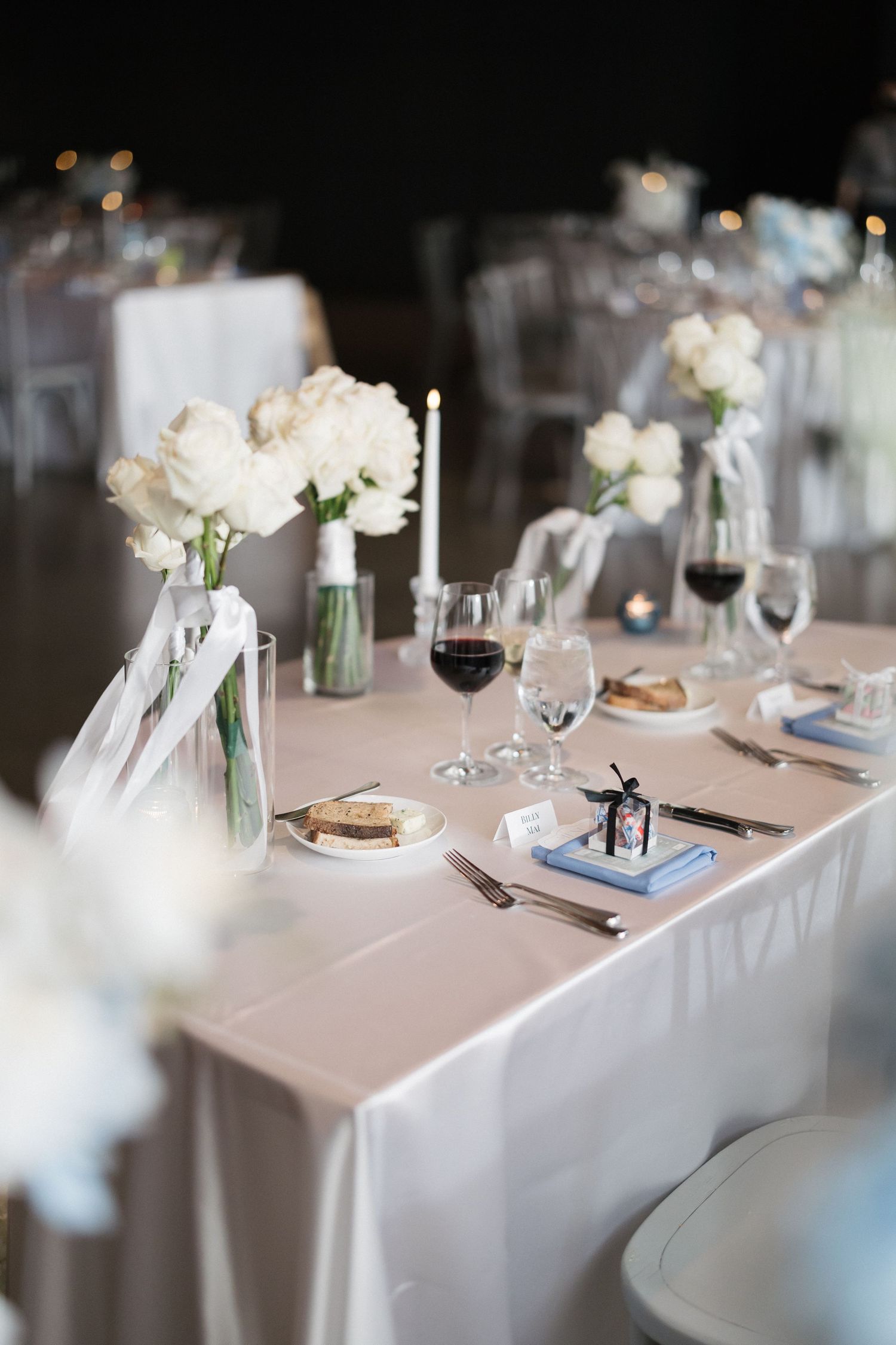 Elegant wedding reception table with white floral centerpieces, candles, red wine glasses, and place settings on a white tablecloth.