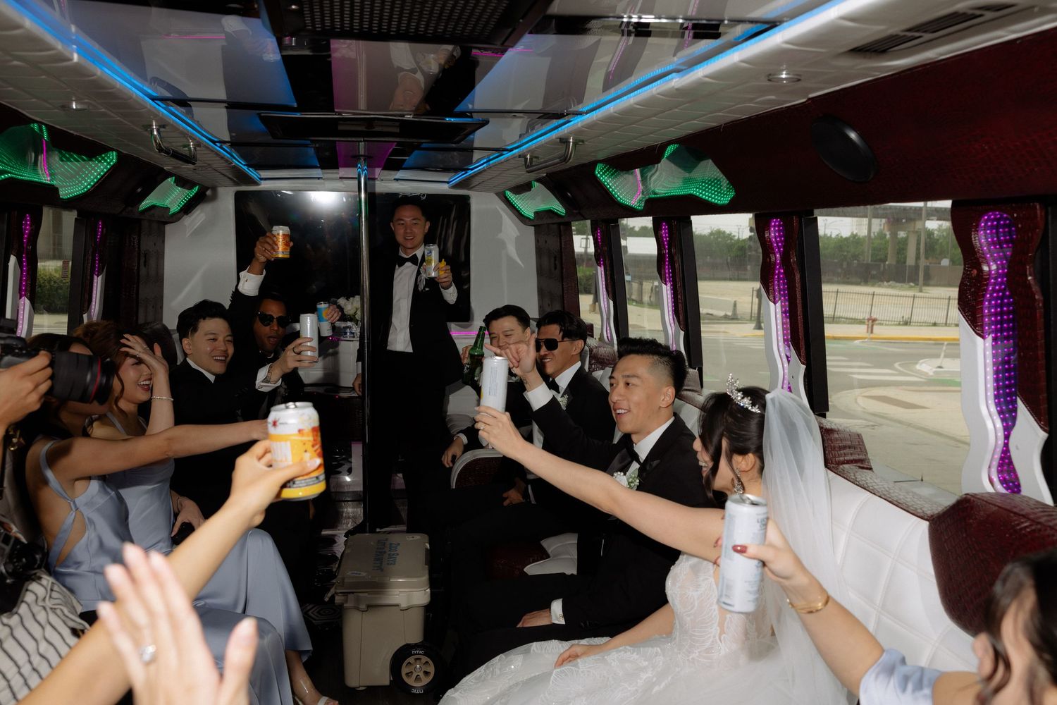 Wedding party toasting with drinks inside a decorated party bus, bride and groom celebrating with guests.