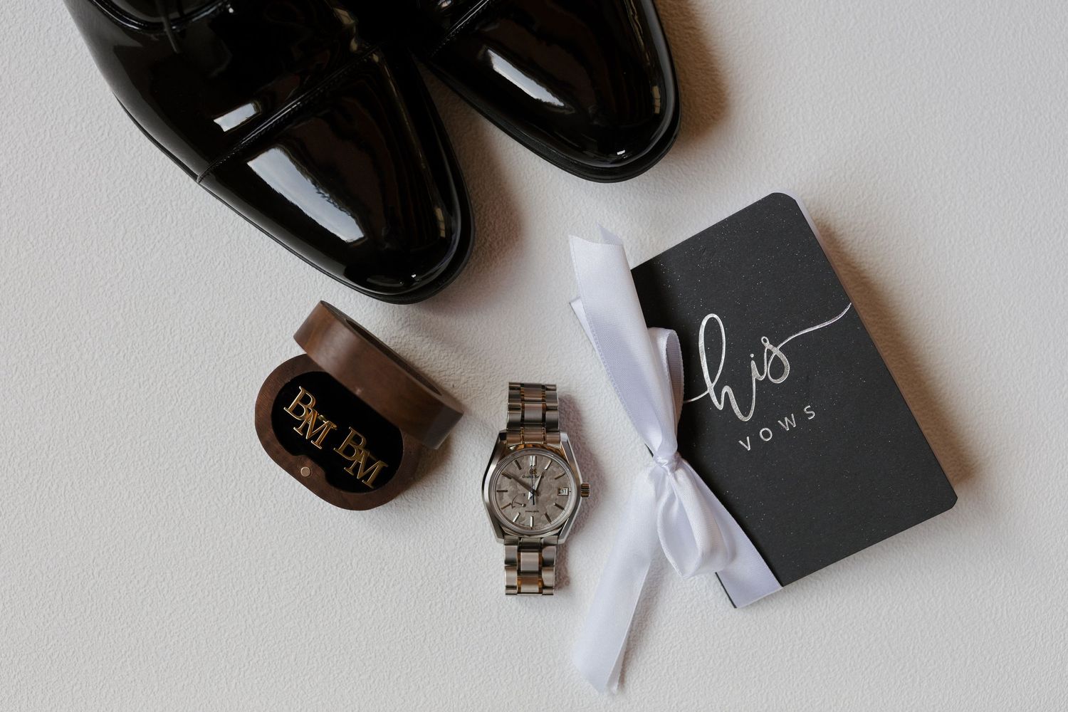 Groom wedding flat lay with black dress shoes, wooden watch, cufflinks, and his vows book on white background.