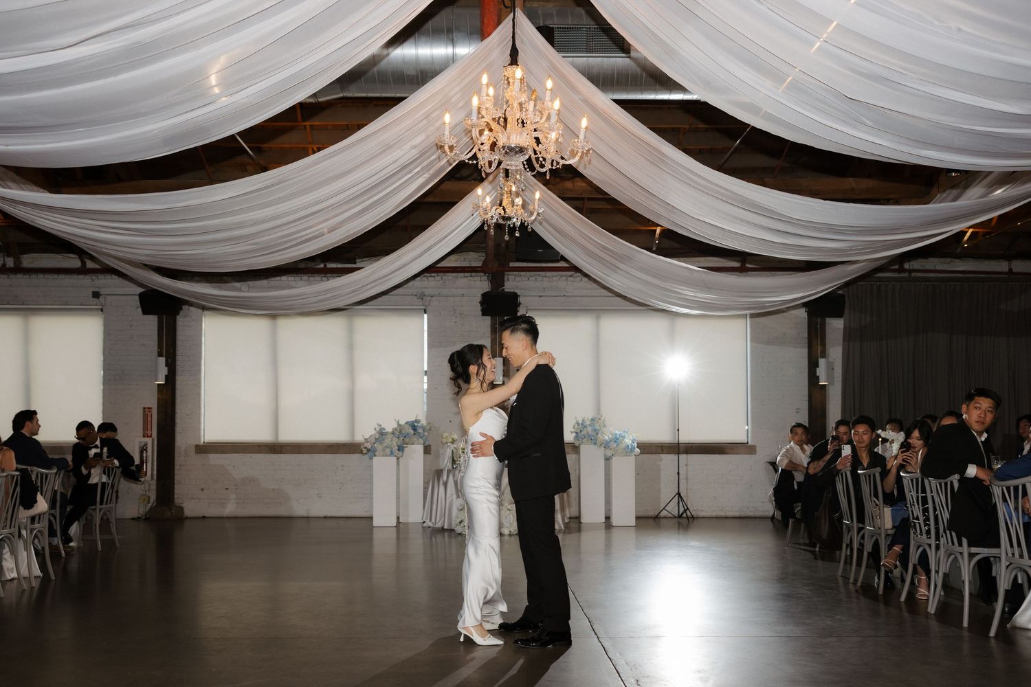 Bride and groom share first dance under elegant white draped ceiling with chandelier at wedding.