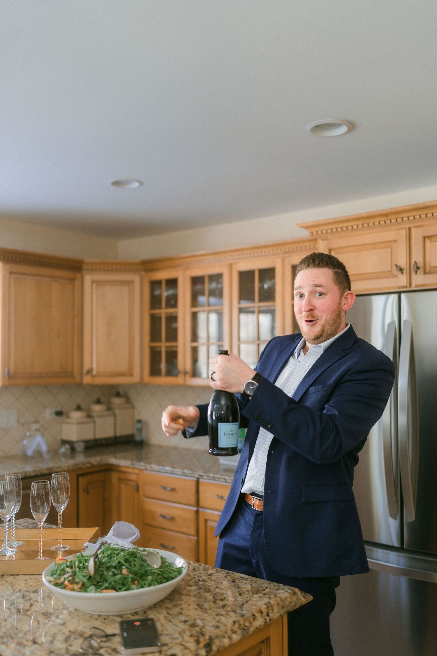 Groom in navy suit opening champagne bottle in kitchen with glasses and salad on granite countertop.
