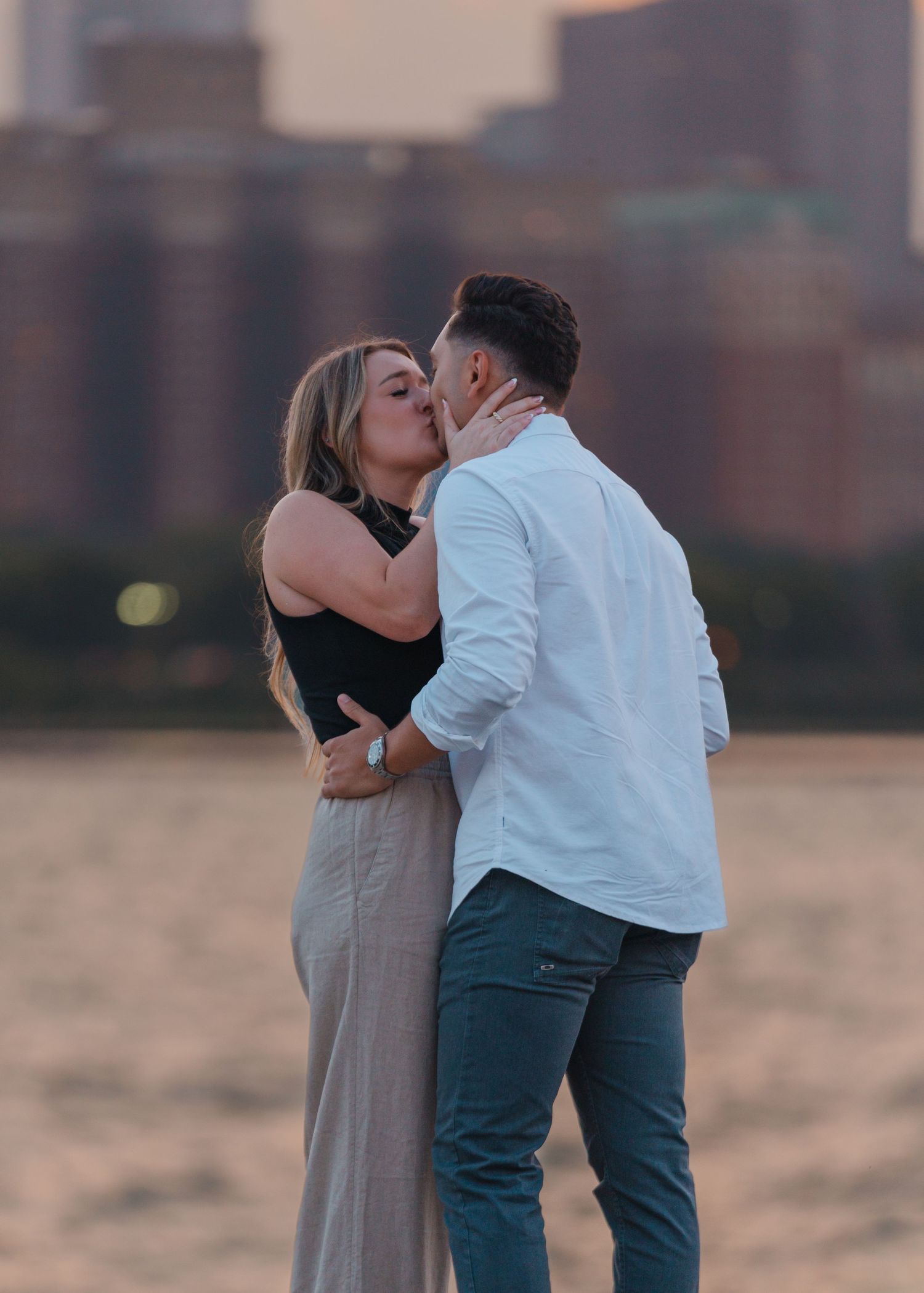 Couple sharing a passionate kiss on a beach with a blurred city skyline at sunset.