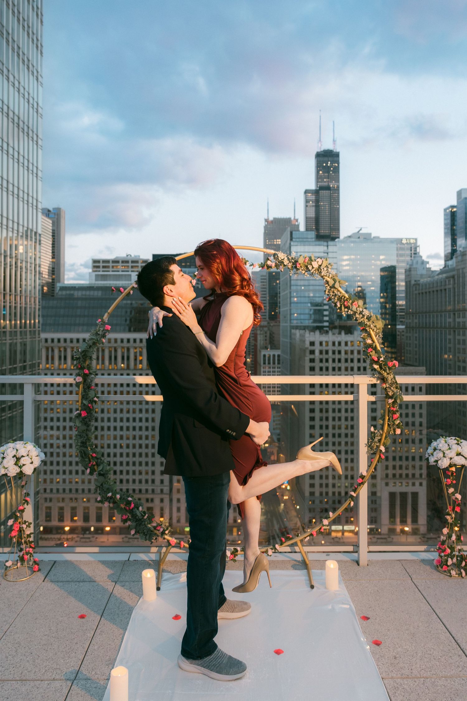 Man lifting woman for a kiss in front of floral hoop arch on Chicago rooftop.