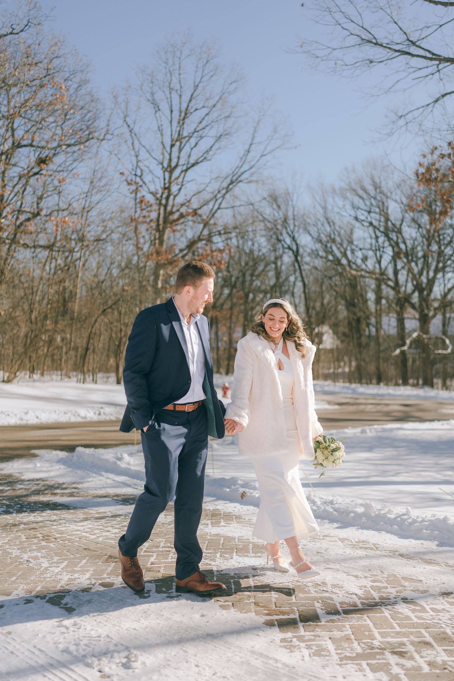 Bride in white fur coat and groom in navy suit walk hand in hand on snowy brick path.