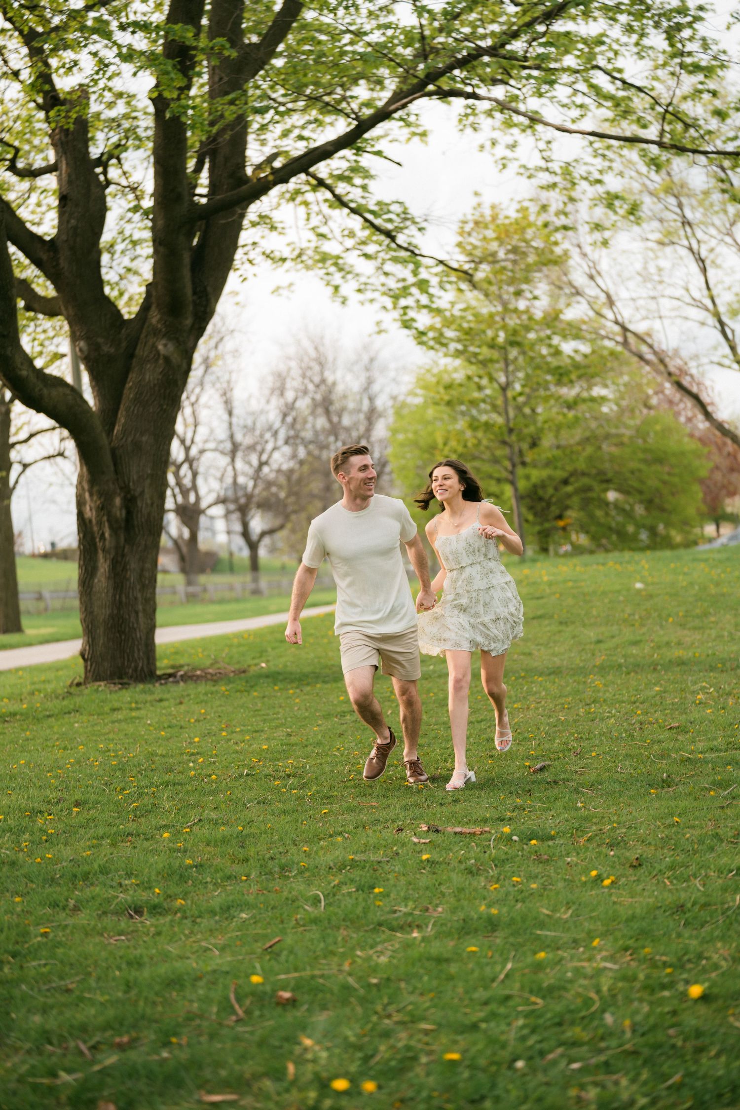 A smiling couple runs hand-in-hand through a green park surrounded by spring trees.