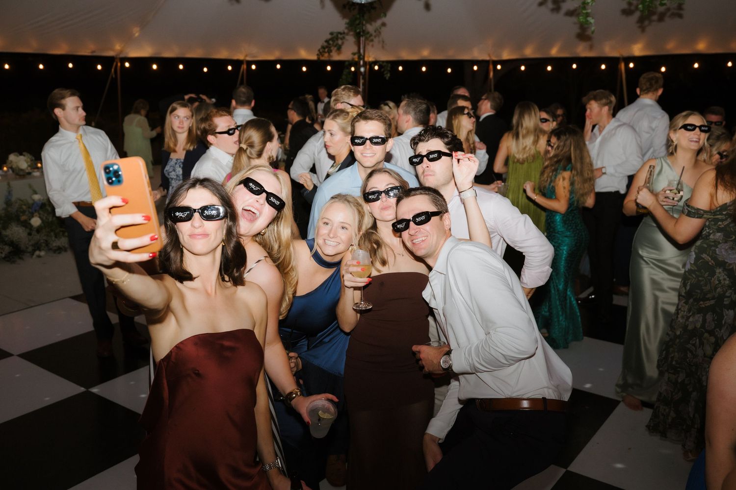 Group of wedding guests pose for fun selfie wearing sunglasses on checkered dance floor at reception.
