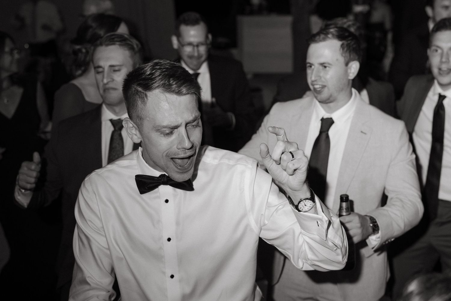 Groom in bow tie dances joyfully with groomsmen at wedding reception in black and white photo.