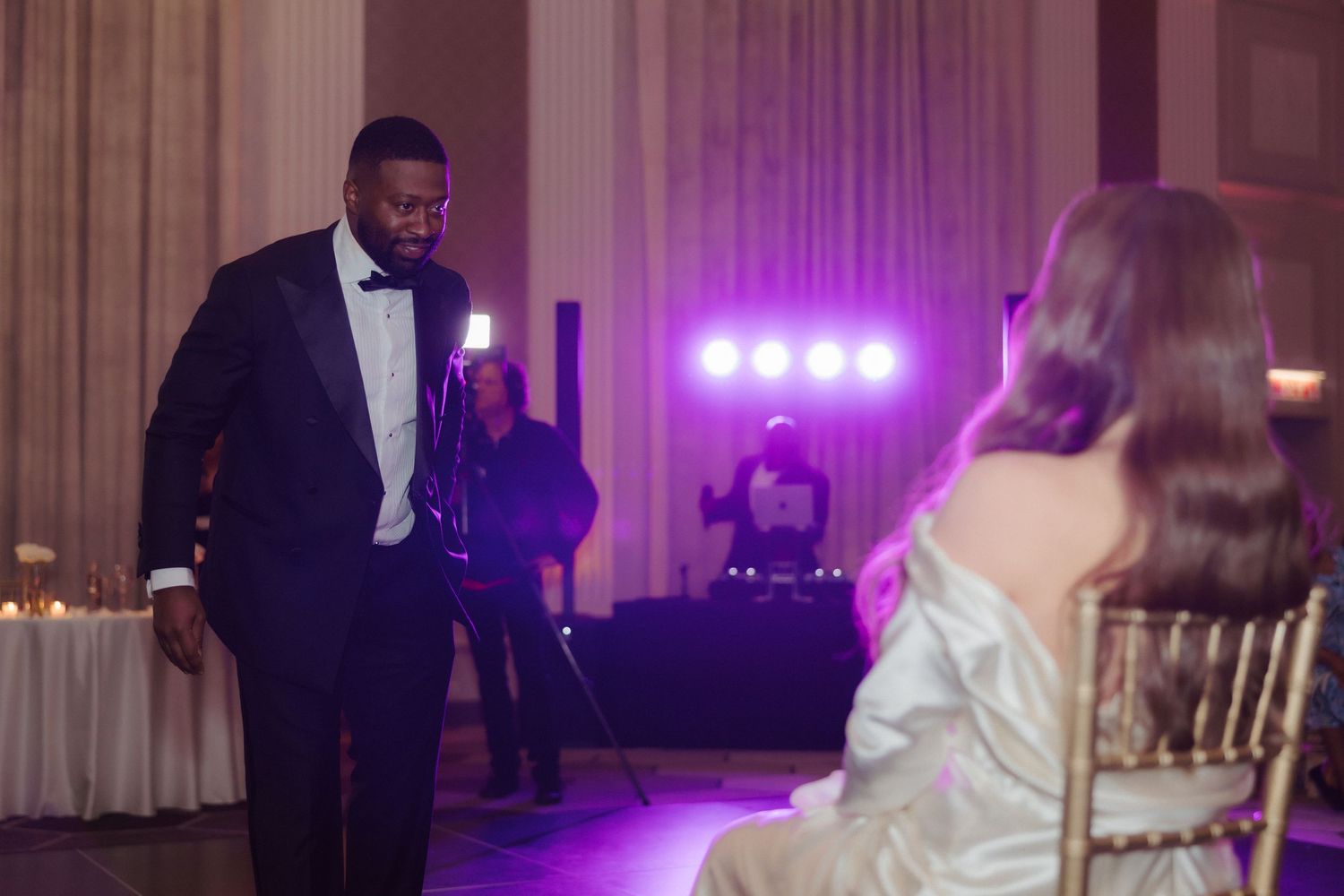 Man in tuxedo bowing to seated woman in elegant gown at formal wedding reception with purple lighting.