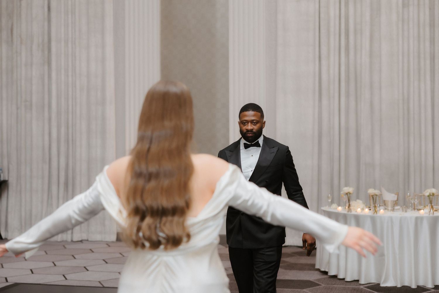 Bride in white gown opens arms to groom in black tuxedo during elegant wedding reception first dance.