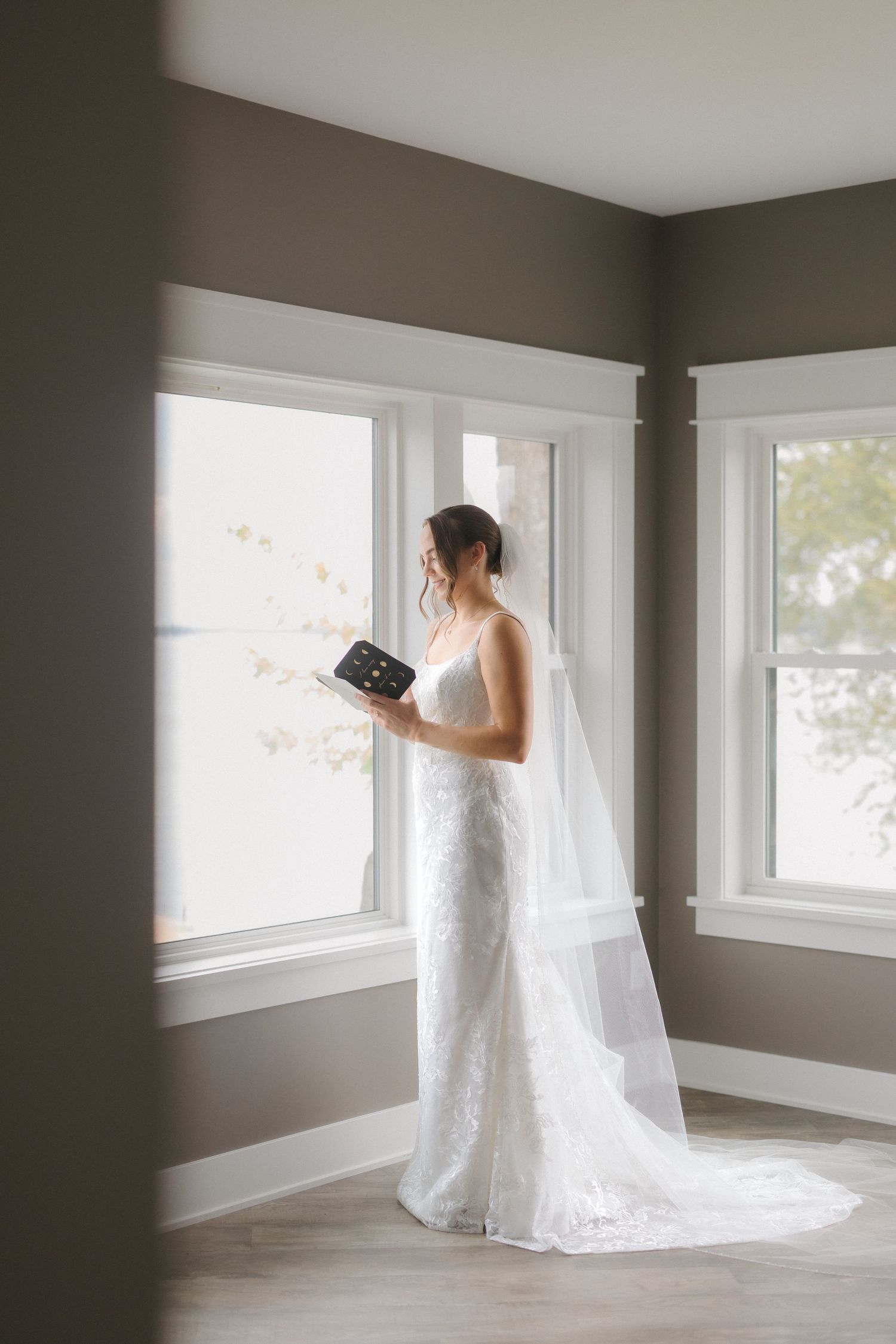 Elegant bride in white lace gown with veil holding a gift box by bright windows.