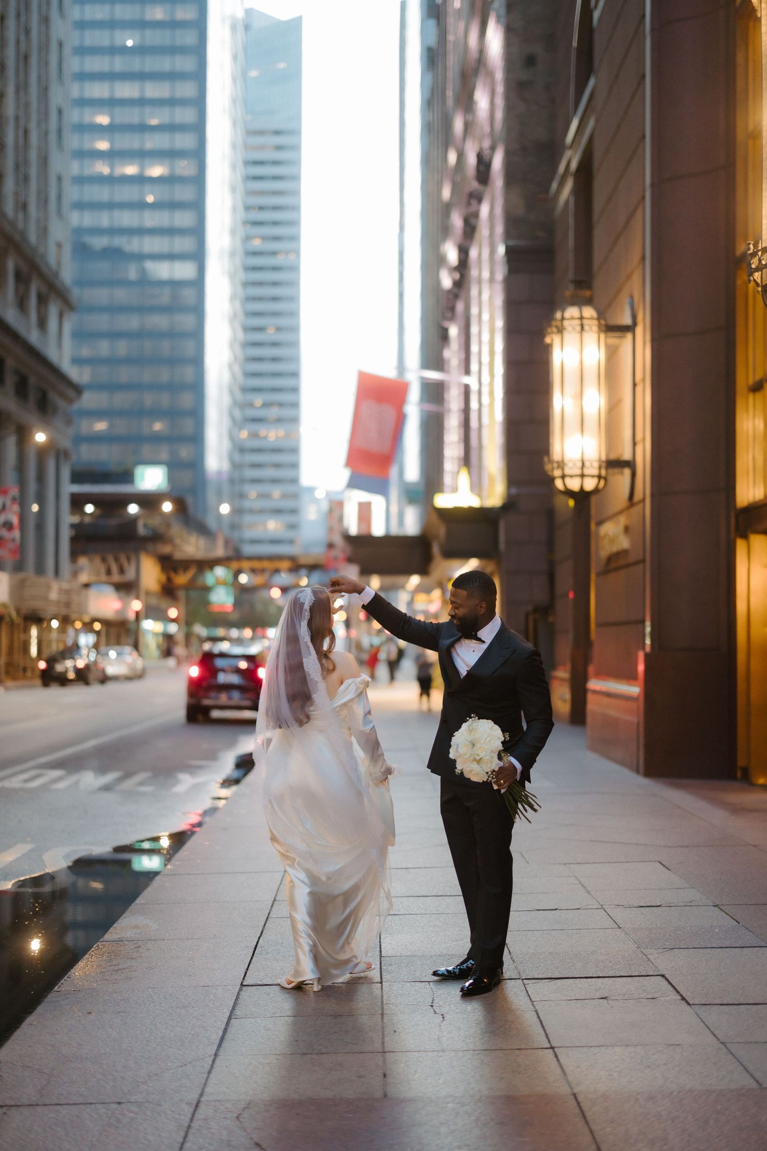 Bride and groom dancing on a city sidewalk surrounded by skyscrapers, groom in tuxedo holding white bouquet.
