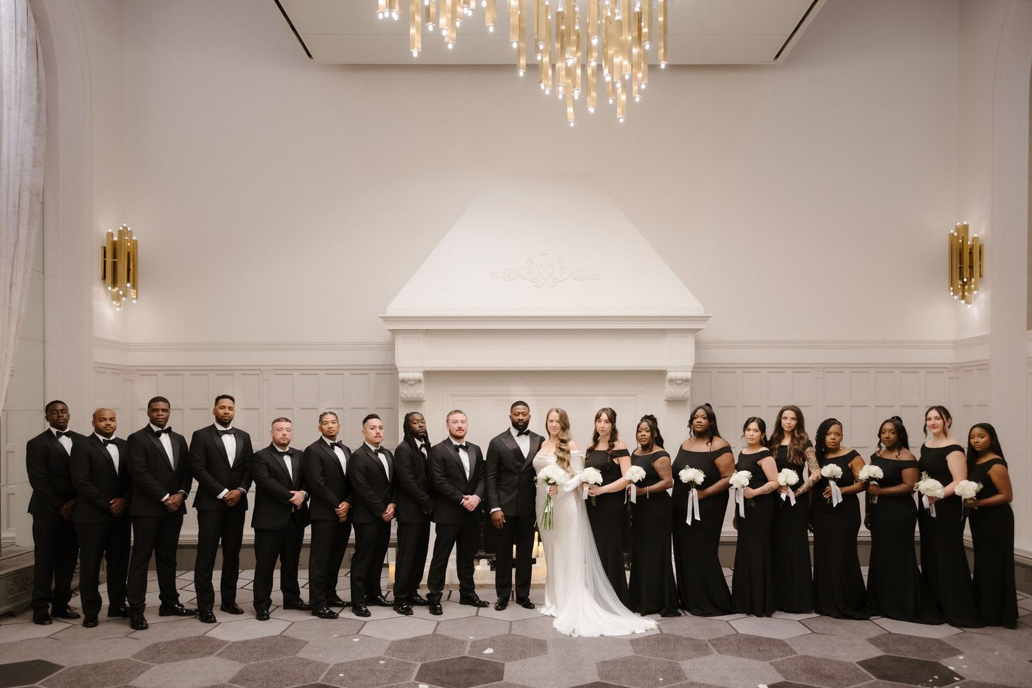 Wedding party in black attire poses in elegant white venue with chandelier, groomsmen and bridesmaids flanking bride and groom.