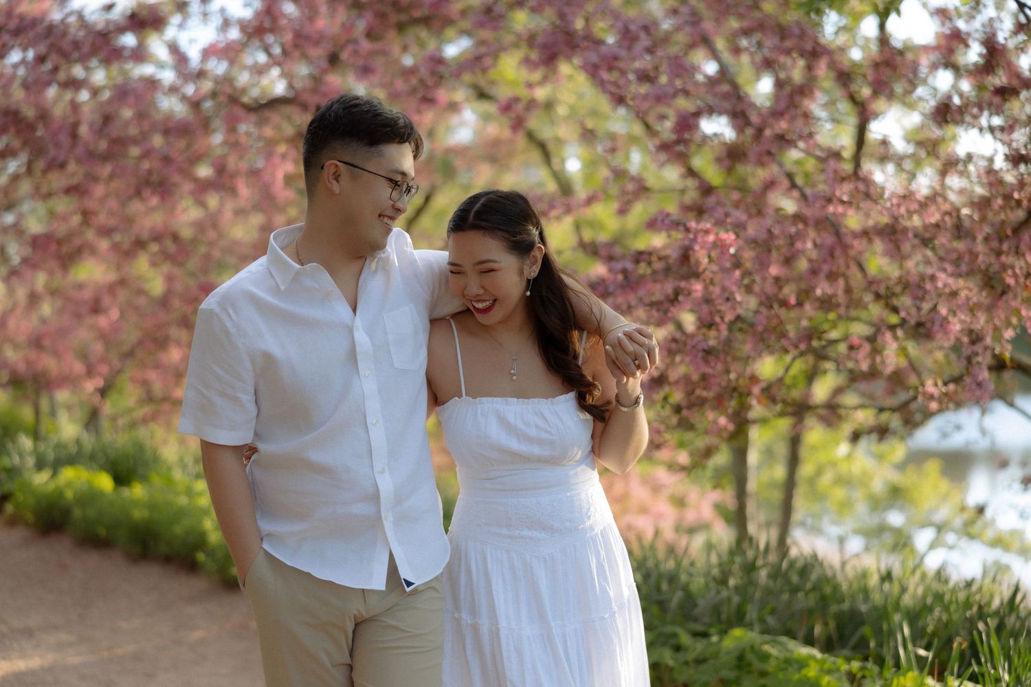 Couple in white outfits embracing under pink cherry blossom trees in a romantic outdoor setting.
