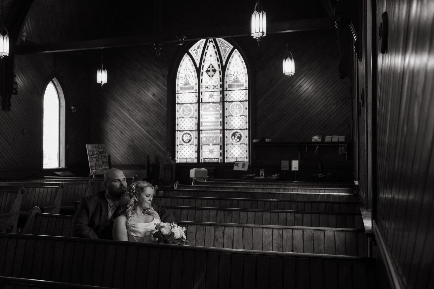 Black and white photo of a couple sitting in a church pew with stained glass windows in the background.