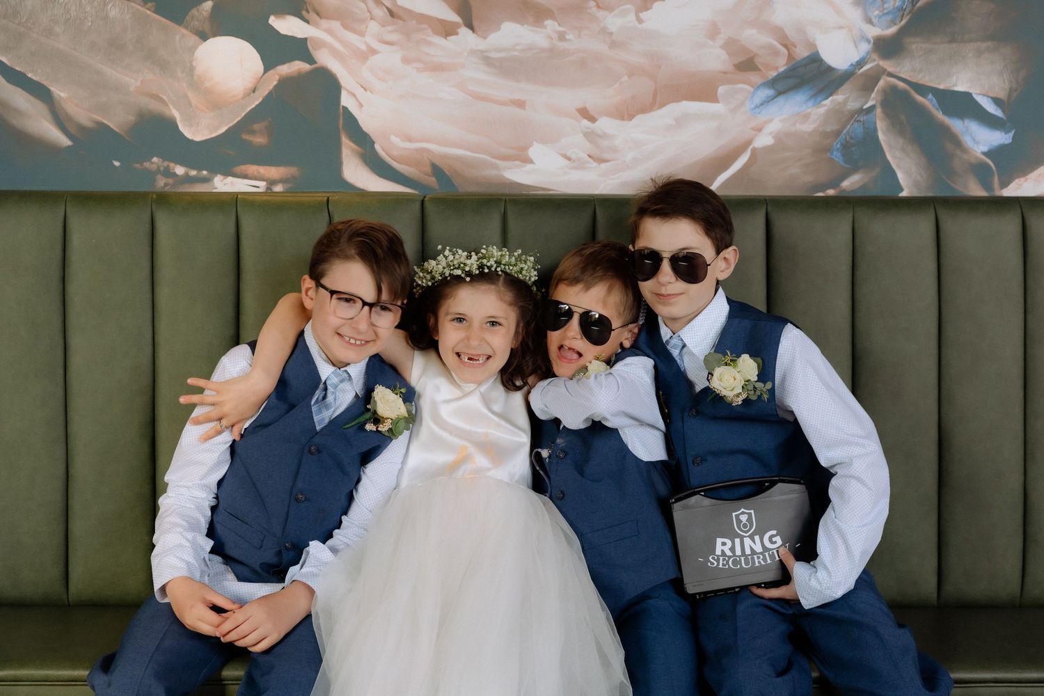 Four children in formal wedding attire pose together on a green velvet bench, smiling and wearing sunglasses.