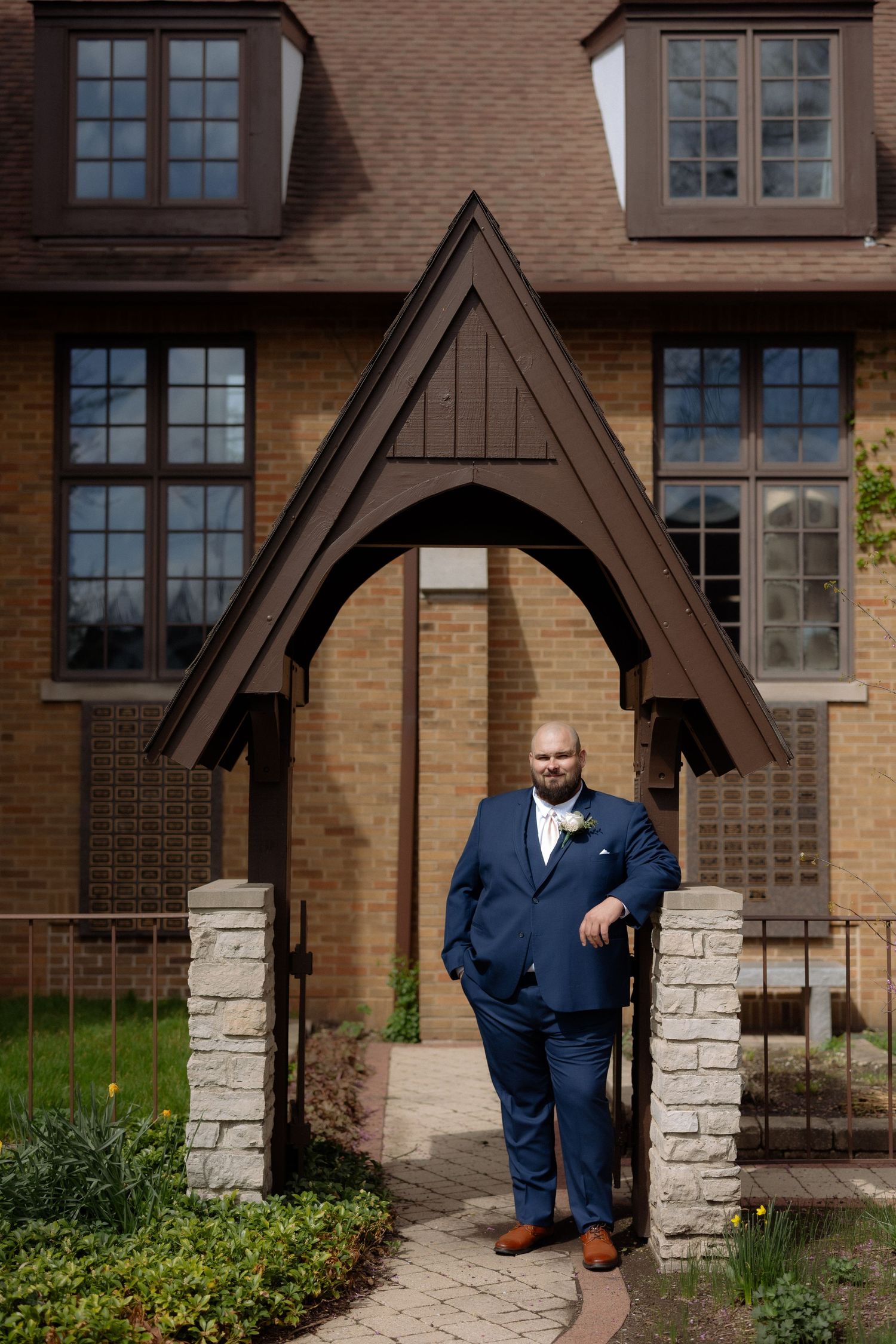 A groom in a navy blue suit with boutonniere poses under a Gothic arch entrance of a brick church.