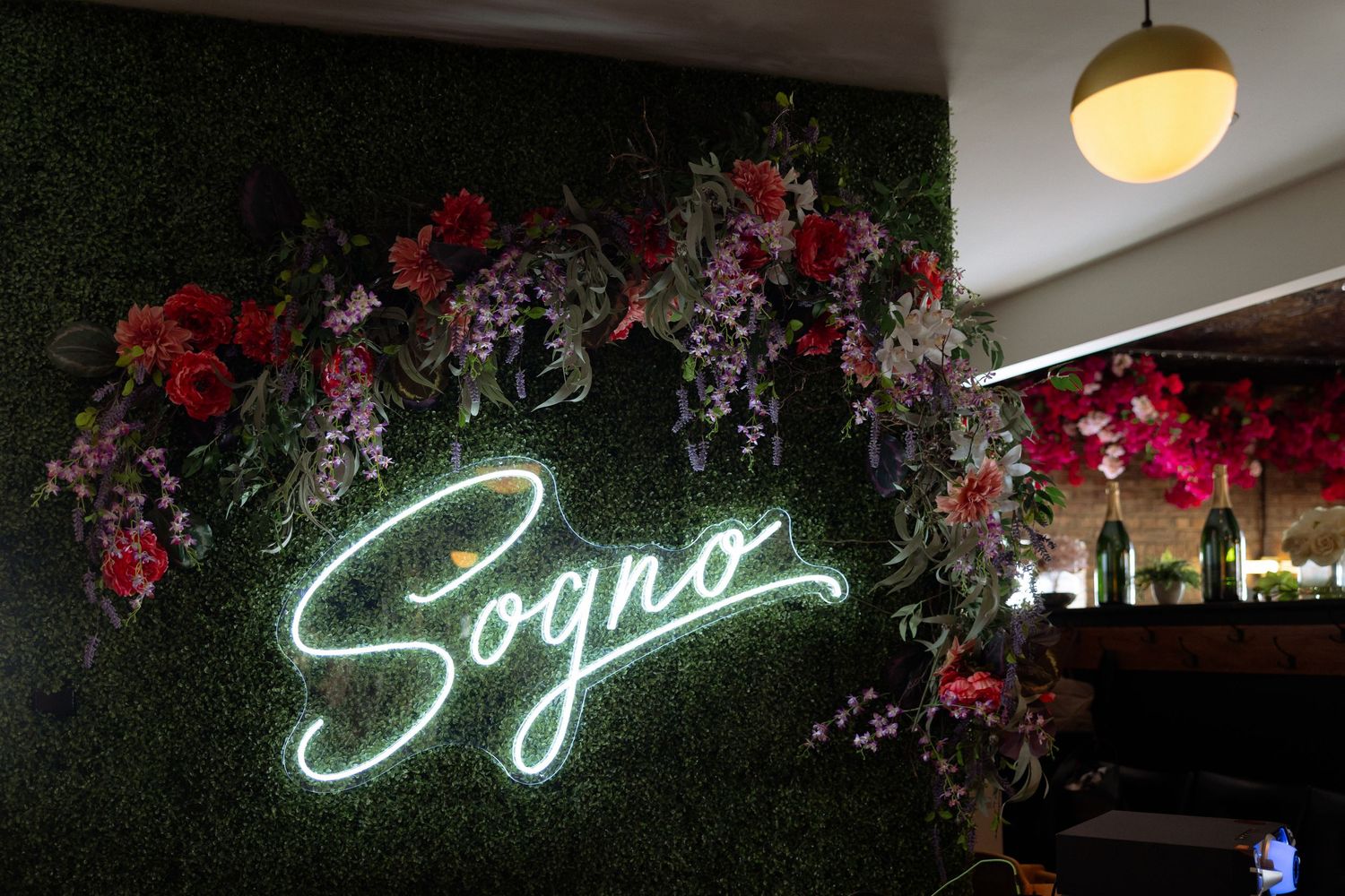 Sogno restaurant neon sign on a lush green wall adorned with colorful artificial flowers.