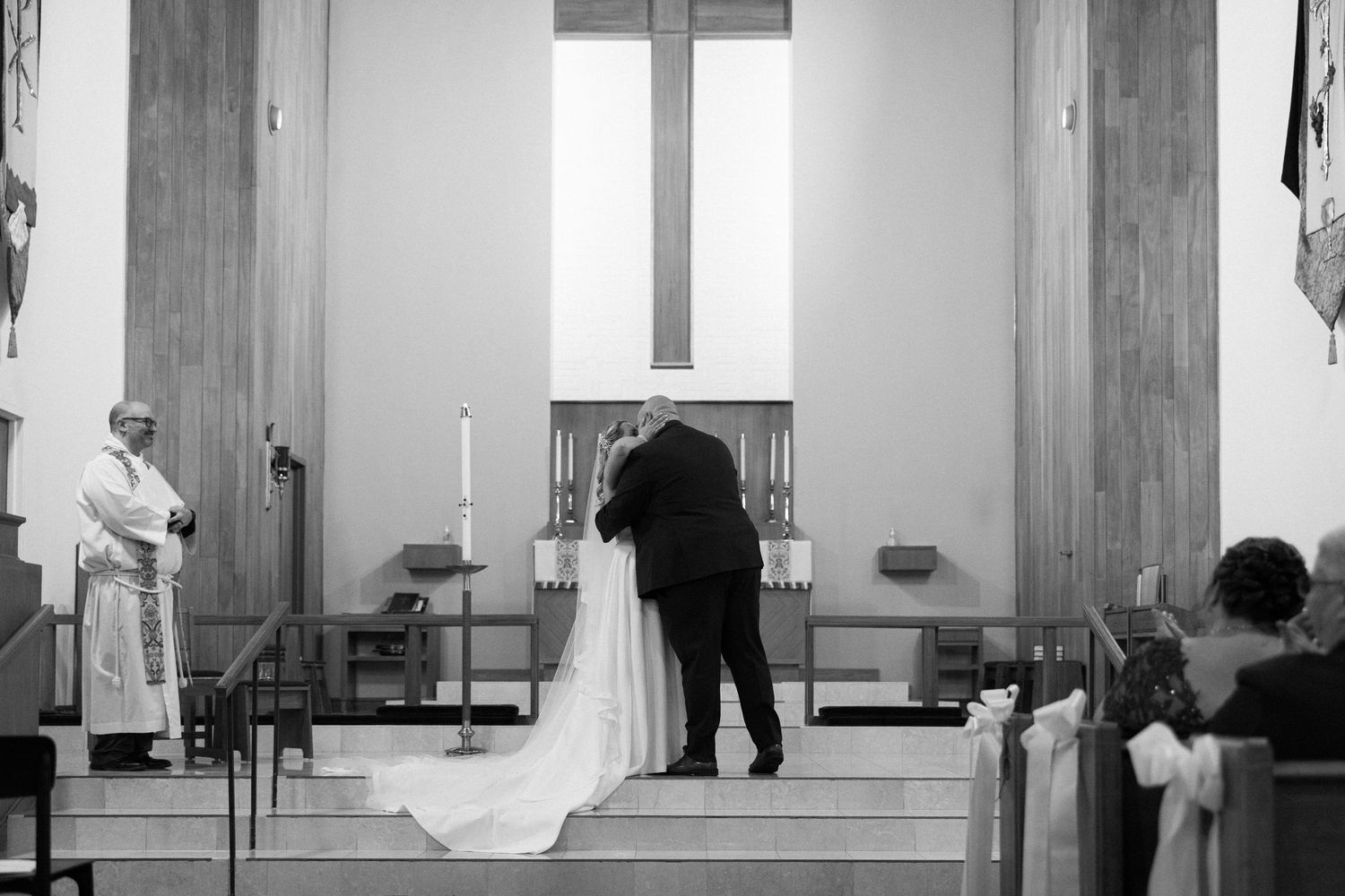 Black and white photo of a couple embracing at a church altar during their wedding ceremony with a priest nearby.