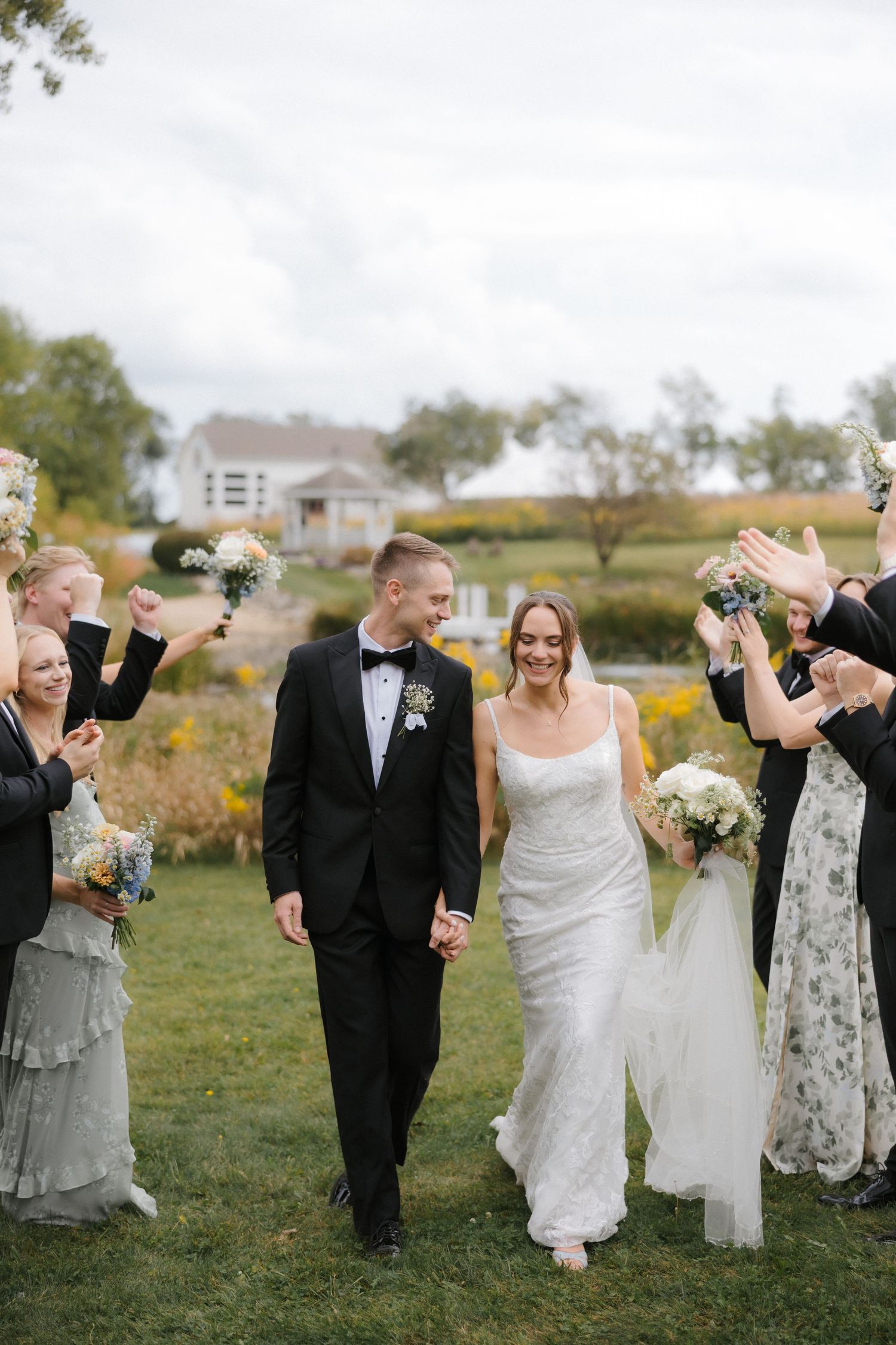 Smiling bride and groom walk hand in hand through cheering wedding party in front of scenic countryside venue.