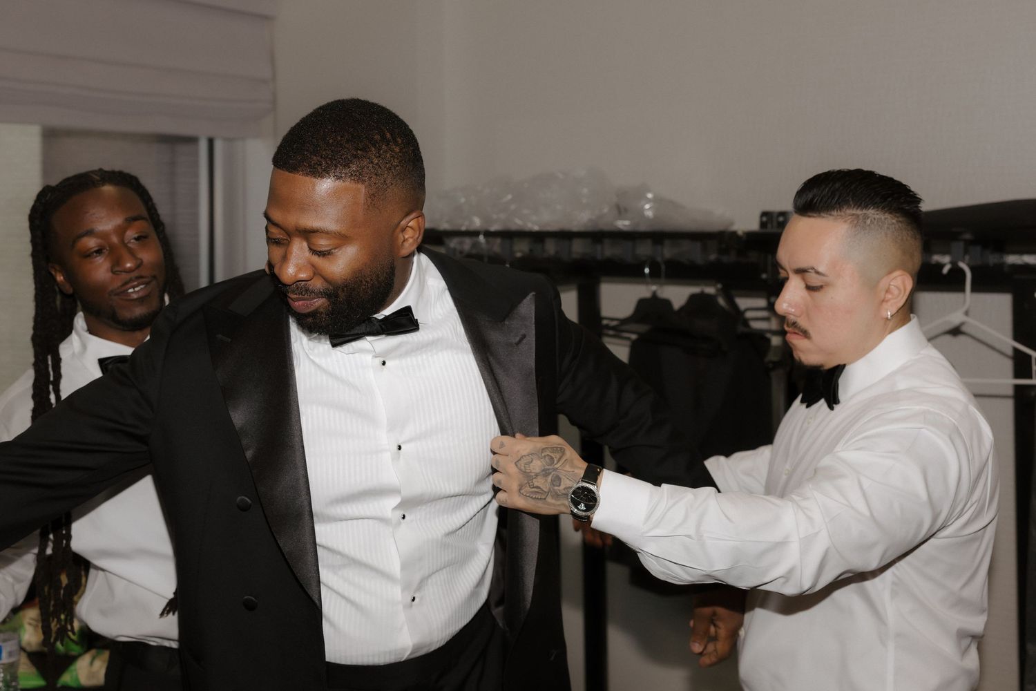 Three men in formal attire, including tuxedos and bow ties, assist each other getting dressed in an elegant room.