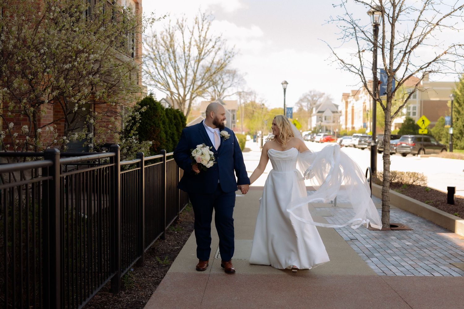Bride and groom walk hand-in-hand on a sunny sidewalk, bride's veil flowing in the breeze, groom in navy suit.