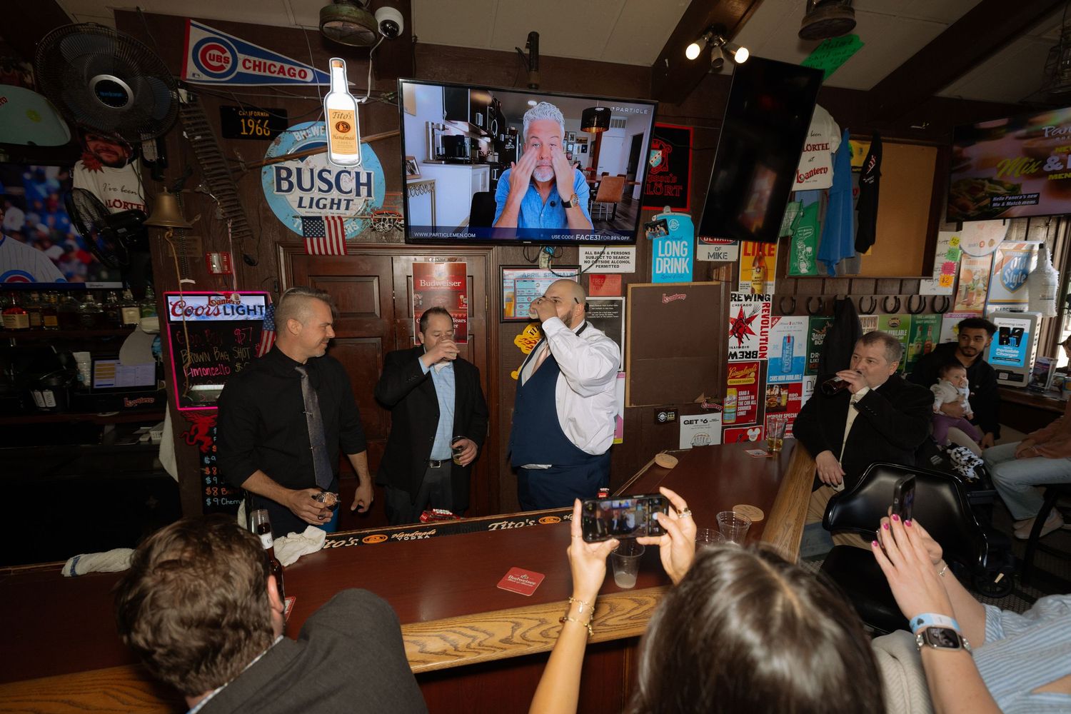 People gather at a bar with Busch Light sign, watching a TV screen while others take photos of the event.