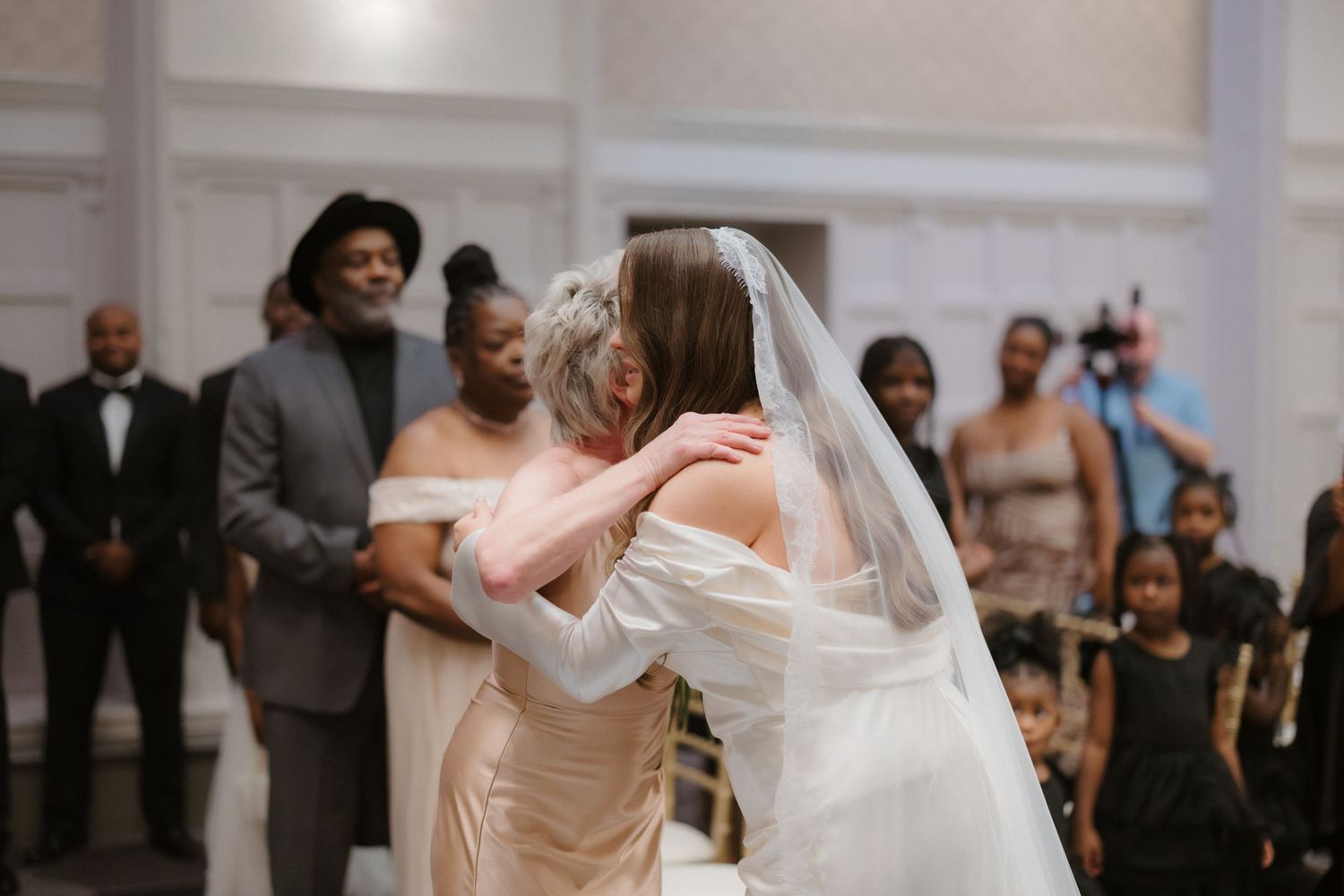 Bride in white gown and veil shares emotional hug with older woman in champagne dress at wedding reception.